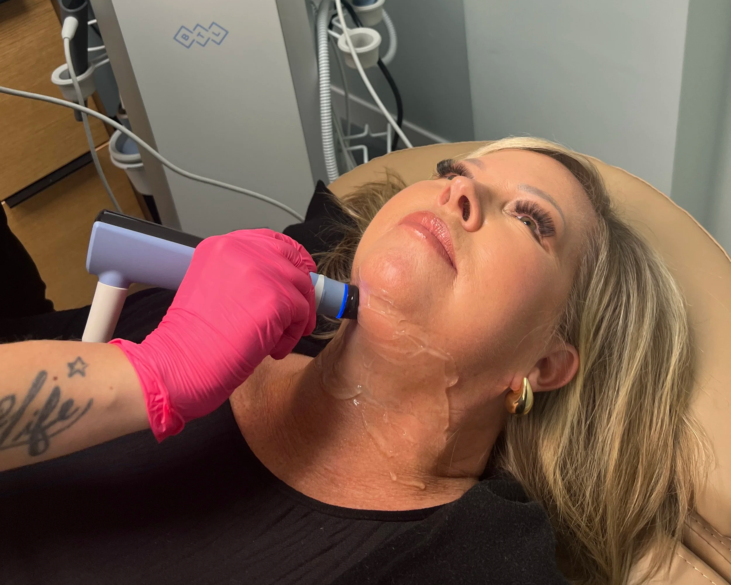 A woman with blonde hair lying on a medical examination table, looking upward, with a healthcare professional in pink gloves examining her face, in a medical setting with medical equipment nearby.