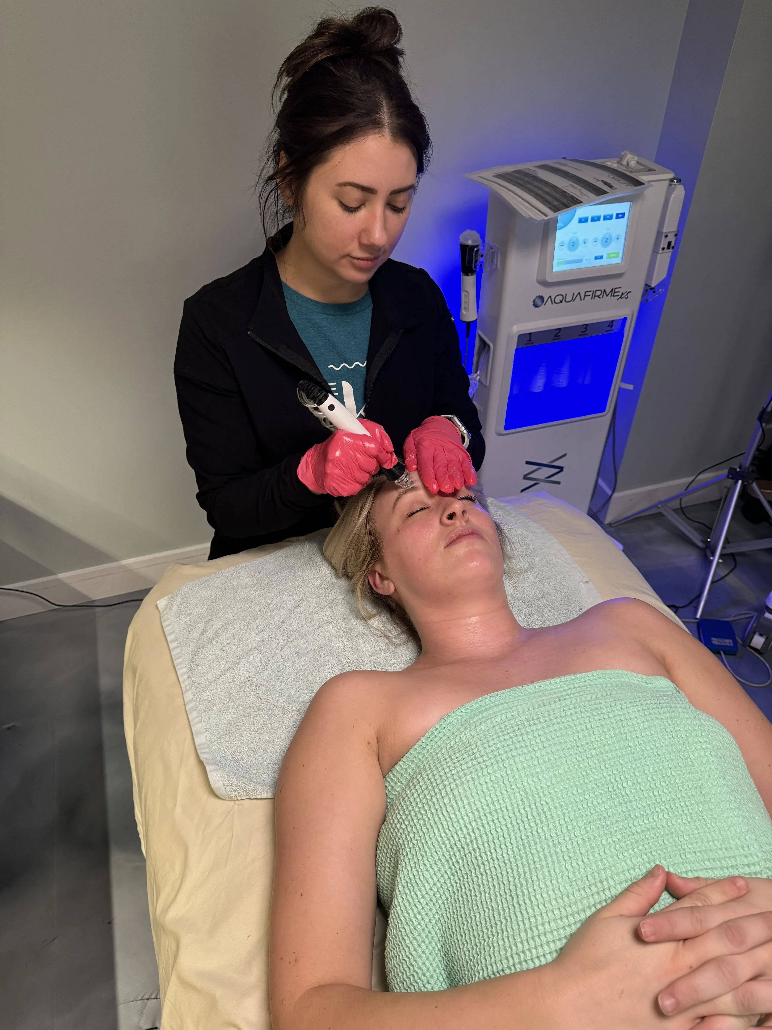 A woman lying on a treatment bed with her eyes closed, receiving a cosmetic facial treatment from a technician who is holding a pen-like device. The technician is wearing pink gloves, and the room has medical equipment and a treatment machine in the background.