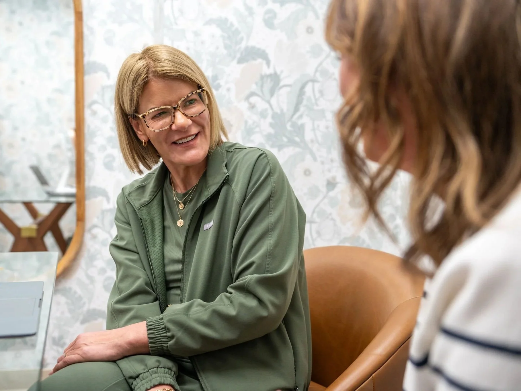 Two women seated in a room, engaged in a conversation. One woman has blonde hair, is wearing glasses, gold jewelry, and a green jacket, smiling and looking at the other woman. The other woman has brown, wavy hair and is wearing a white top with black stripes, but her face is partially out of the frame.