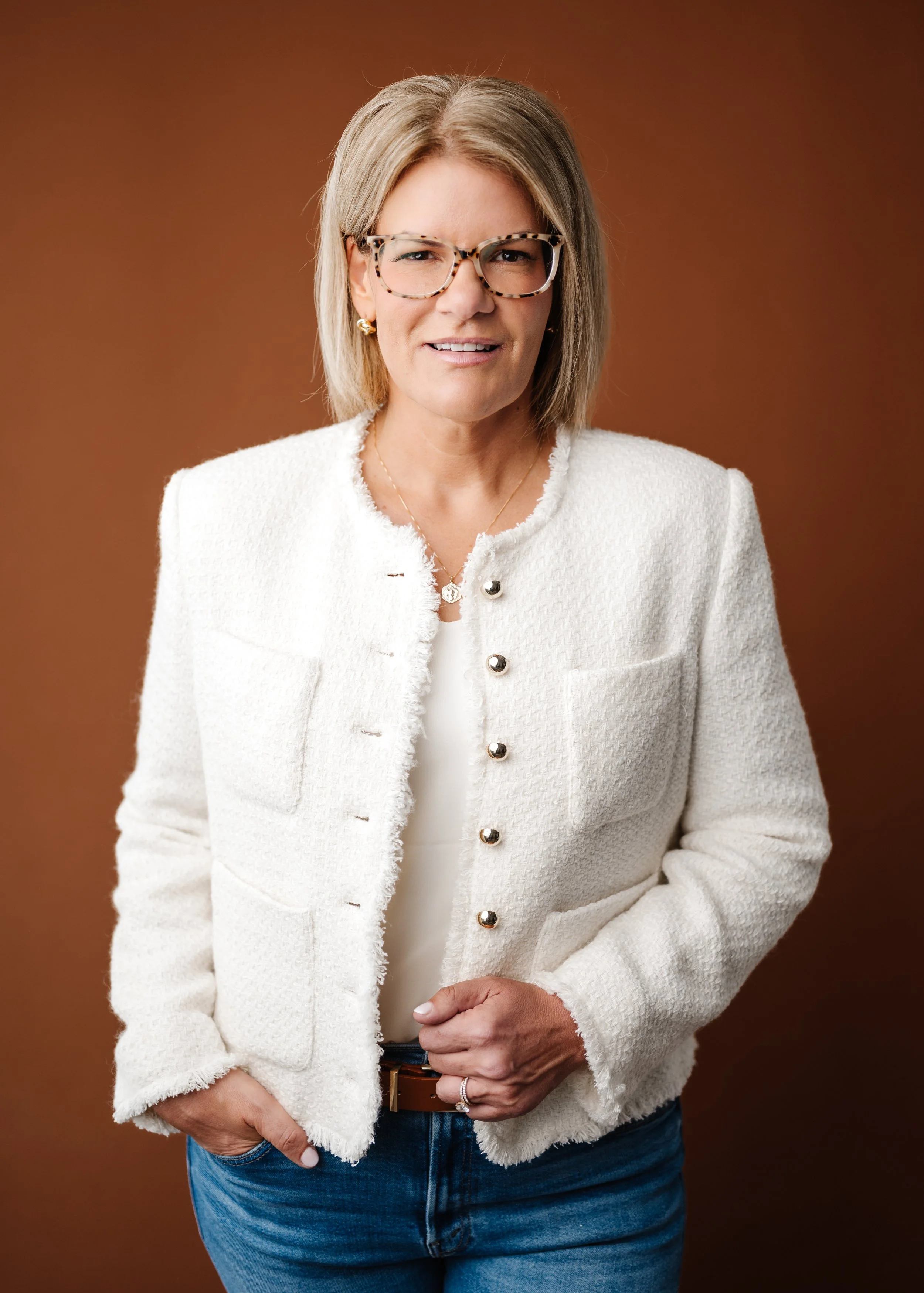 A middle-aged woman wearing glasses with tortoiseshell frames, a white textured blazer with metallic buttons, gold earrings, a necklace, and a ring on her finger, standing against a brown background.