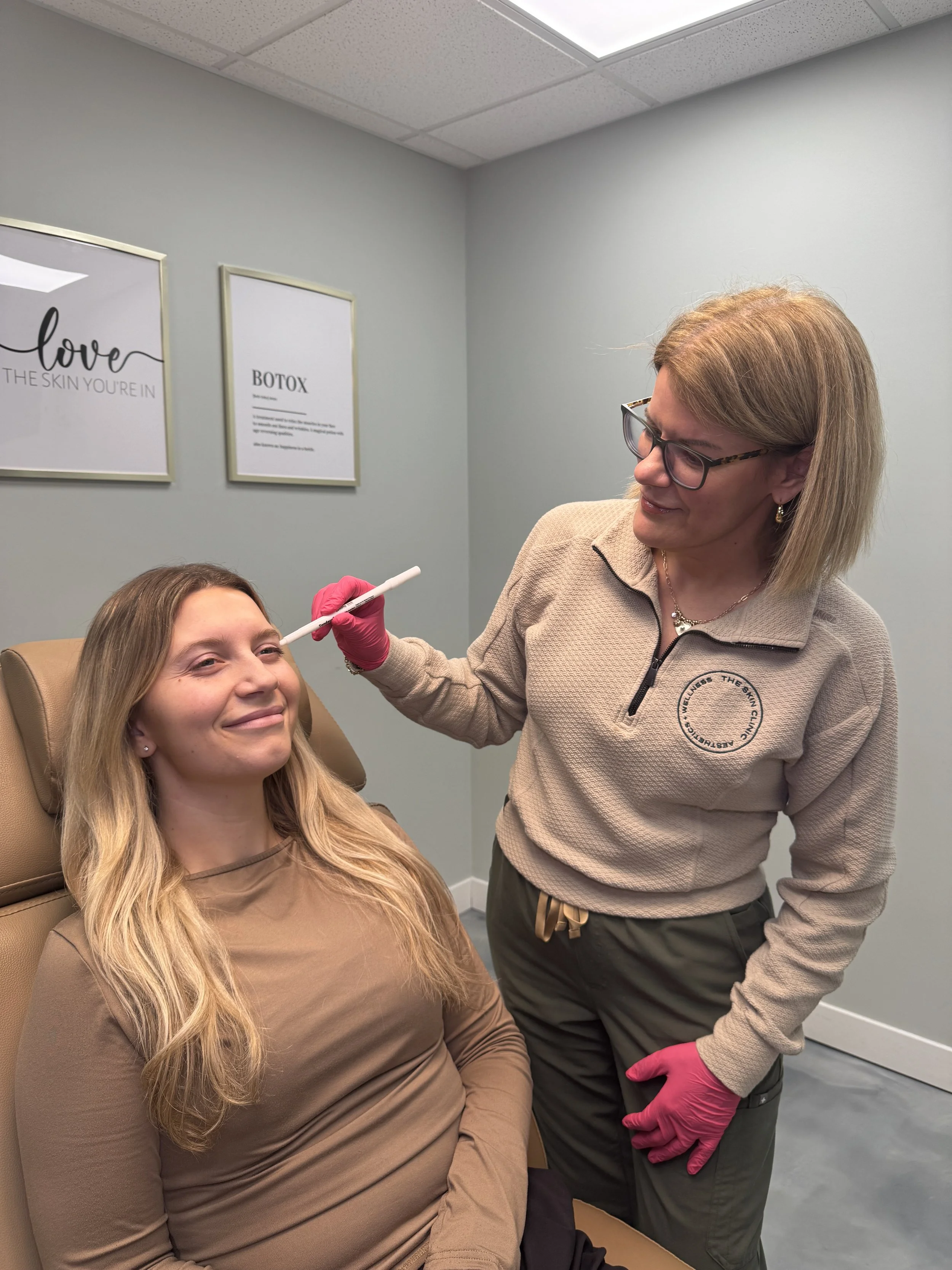 A woman receiving Botox injections from a healthcare professional in a medical office. The woman is sitting in a chair, smiling, with her eyes closed as the healthcare professional, wearing glasses and pink gloves, administers the injection to her face.