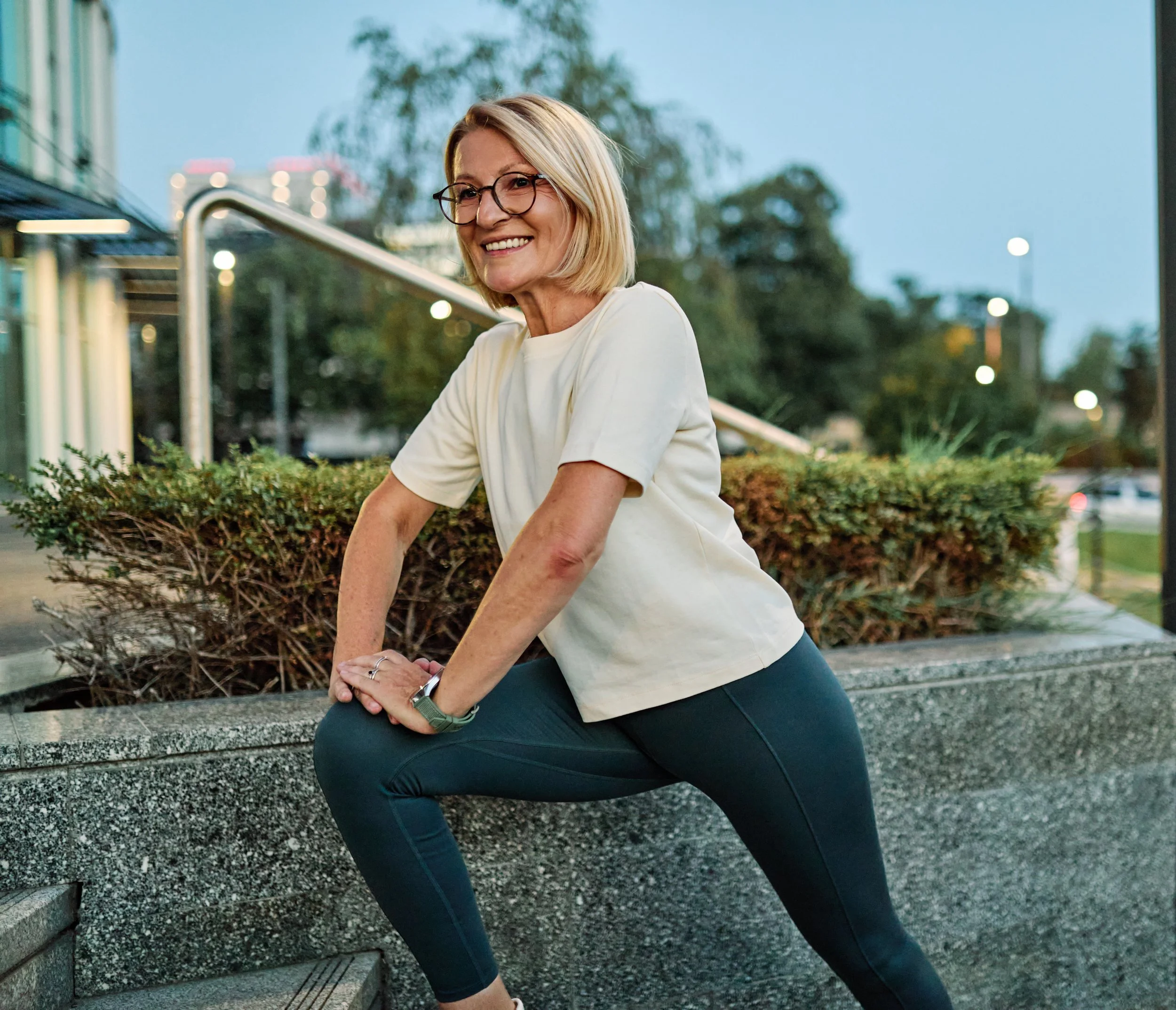 Woman doing a stretching exercise outdoors near a concrete ledge with bushes, buildings, and trees in the background during twilight.