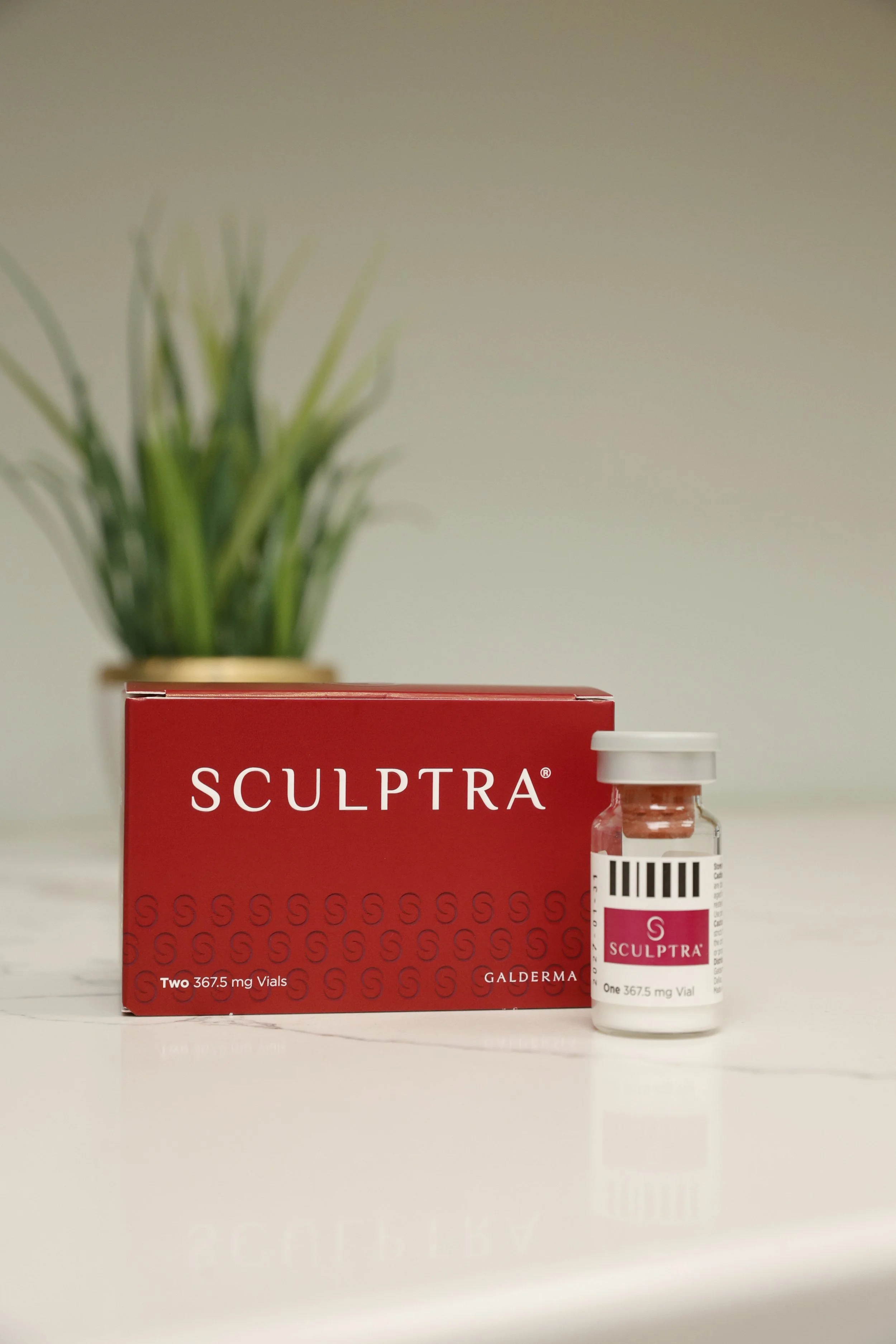 A box and a vial of Sculptra, a cosmetic injectable, placed on a white surface with a blurred green plant in the background.
