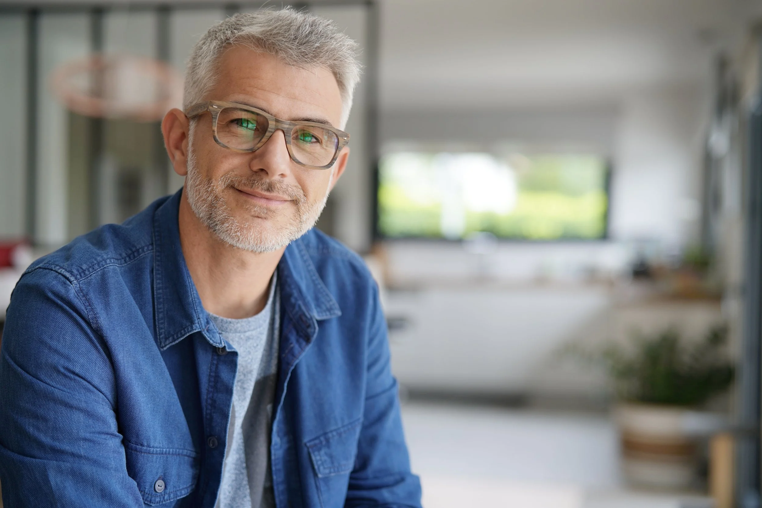 A smiling older man with gray hair, glasses, and a beard, wearing a denim jacket, sitting in a bright, modern indoor space.