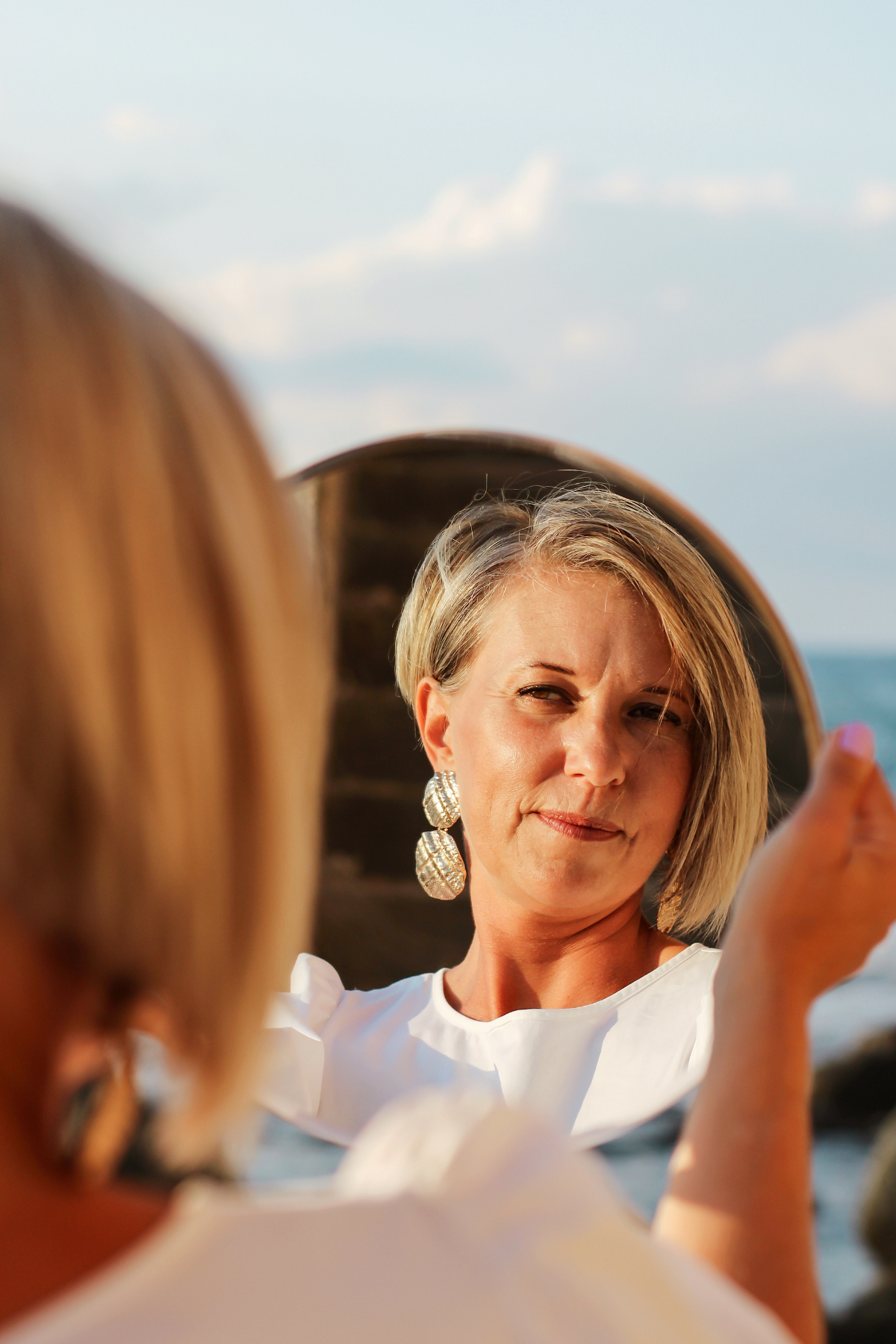 A woman with short blonde hair and large silver earrings is looking at herself in a mirror, holding a phone or camera to take a selfie outdoors by the water during the daytime.