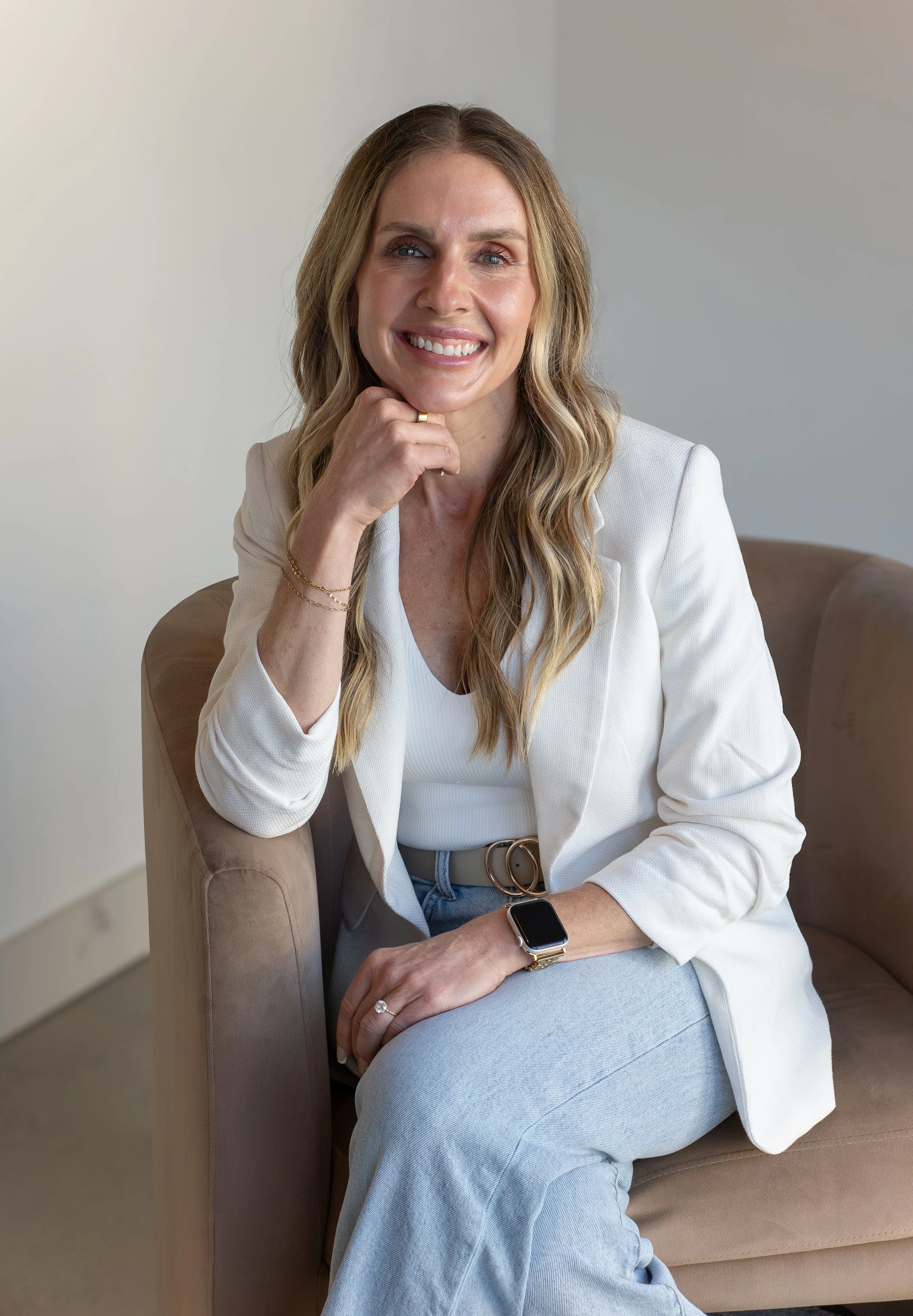 A woman with long wavy blonde hair sitting on a beige couch, wearing a white blazer, white top, light blue jeans, and a smartwatch, smiling at the camera.