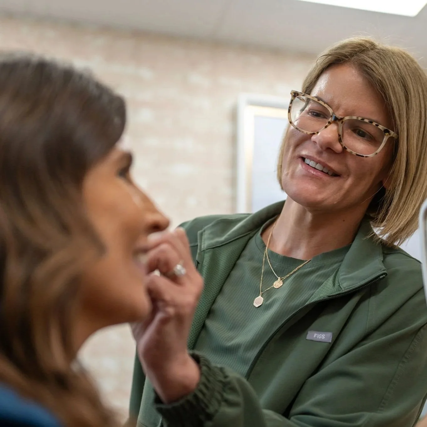 A woman with glasses and blonde hair smiling as she touches a woman's face during a conversation in an indoor setting.