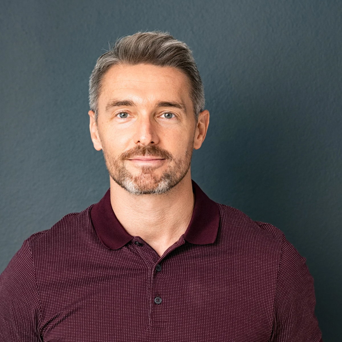 A man with light skin, short grayish hair, and a beard, wearing a maroon polo shirt, standing against a dark teal background.