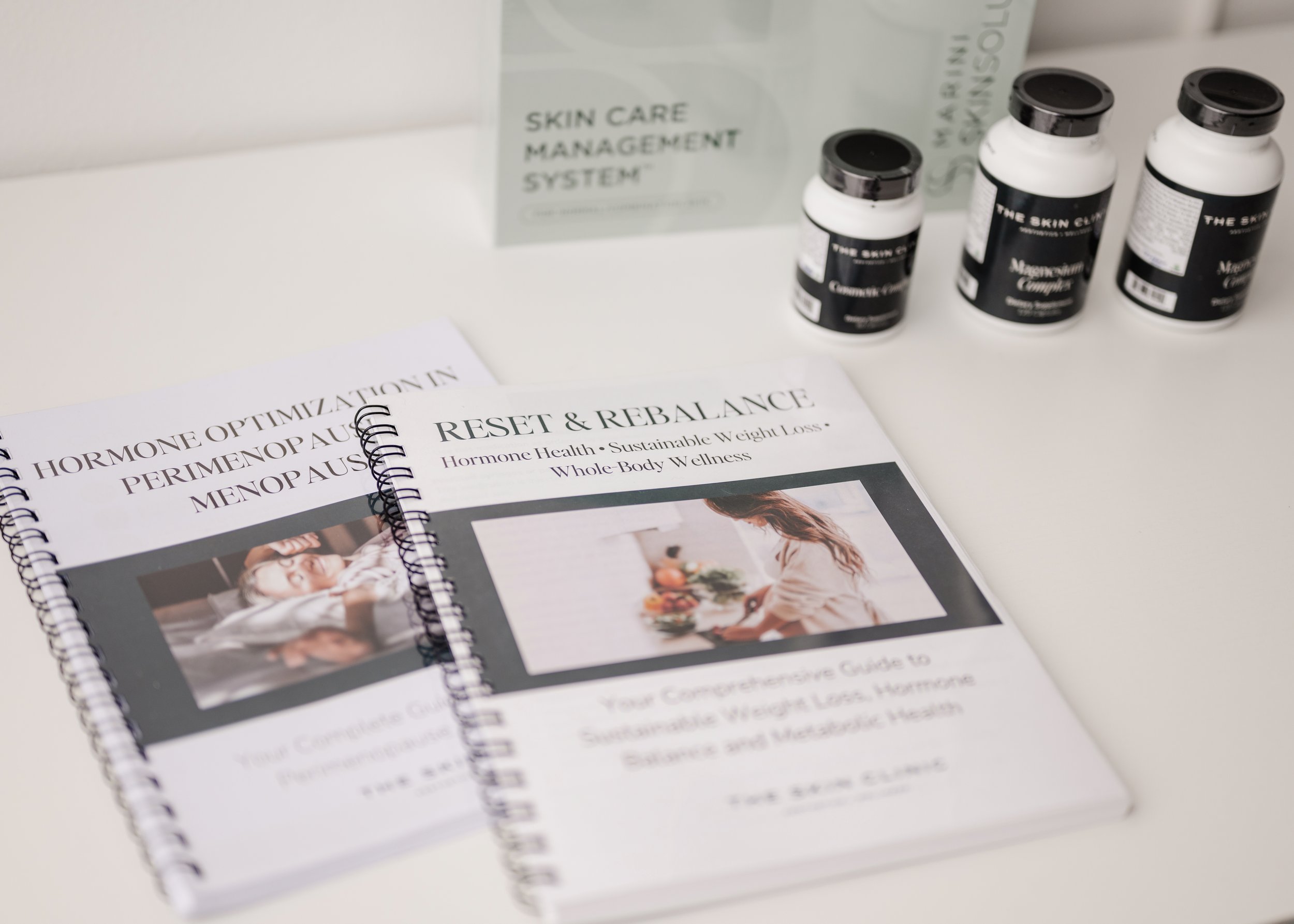 A white surface with a booklet titled 'Hormone Optimization in Perimenopause and Menopause' and a planner titled 'Reset & Rebalance' beside four white bottles with black caps labeled 'The Skin Camp' and 'Maximum Complex'.
