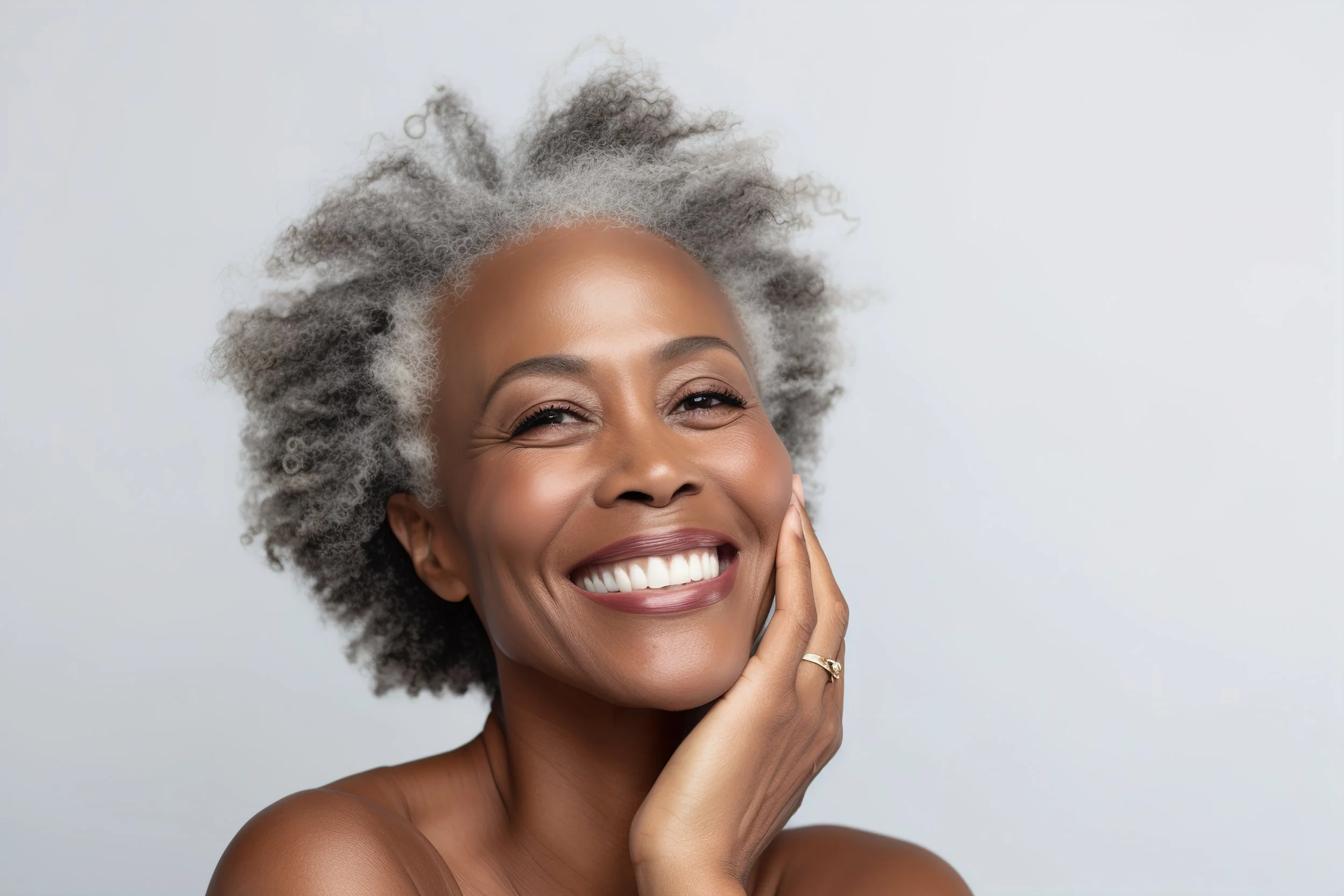 A smiling mature woman with gray, curly hair, touching her face with one hand, showing her white teeth and smooth skin.