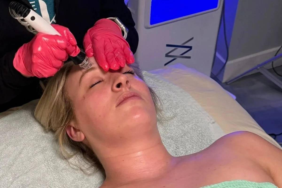 A woman lying on a treatment bed with her eyes closed, receiving a cosmetic facial treatment from a technician who is holding a pen-like device. The technician is wearing pink gloves, and the room has medical equipment and a treatment machine in the background.