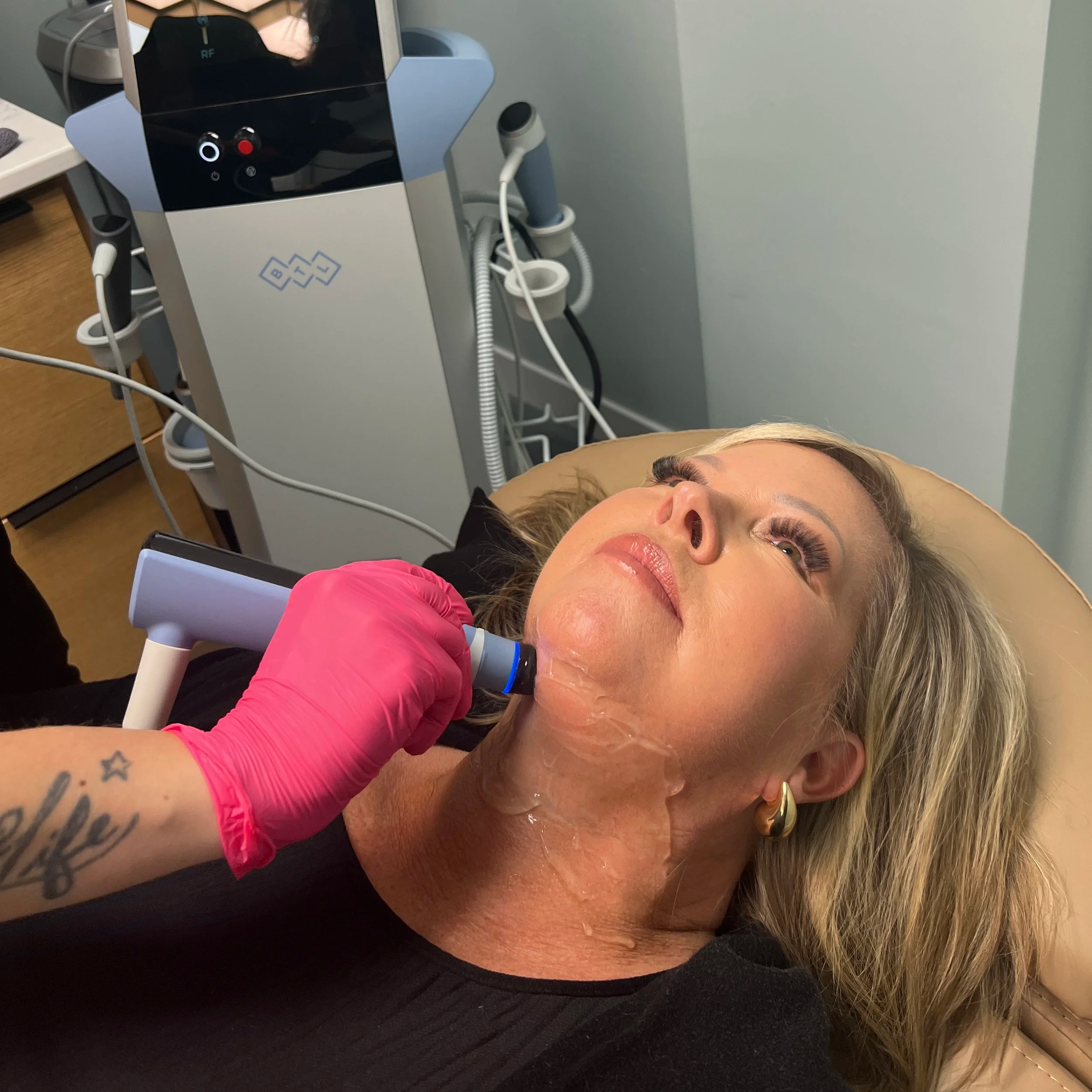 A woman with long blonde hair lying on her side in a medical setting, possibly a clinic or hospital, with a medical device nearby and her arm visible with tattoos. She is wearing a pink garment.