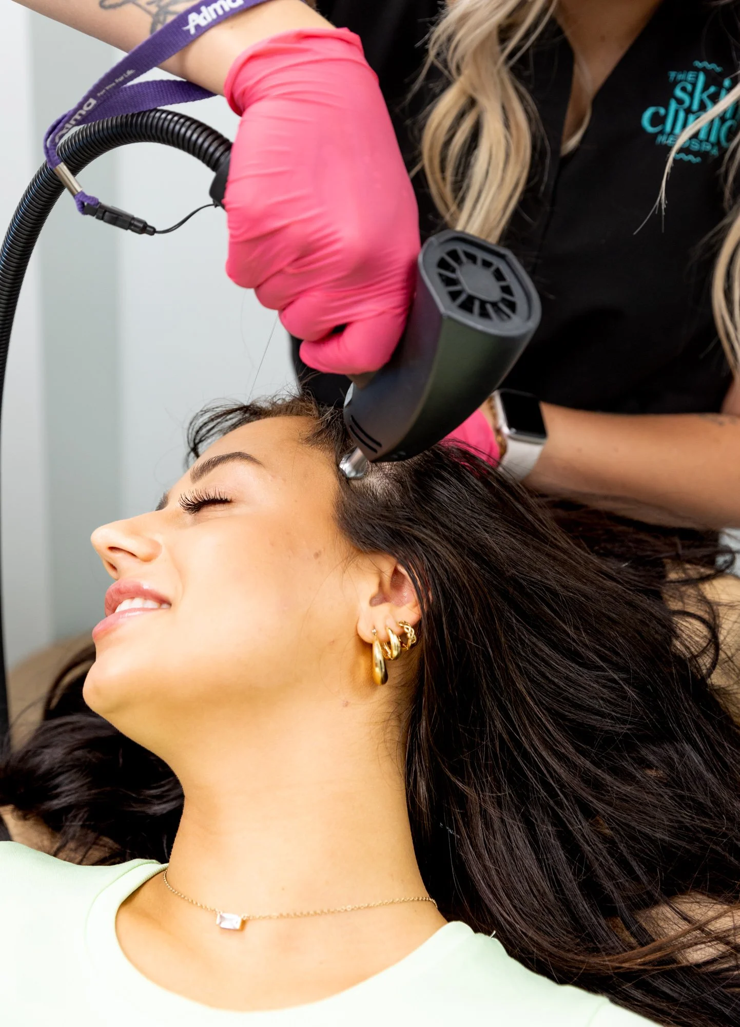 A woman receiving a cosmetic treatment with a device on her temple, lying down with eyes closed, wearing gold hoop earrings, a light green top, and a thin necklace, while a technician in a black shirt with a logo and pink gloves operates the device.