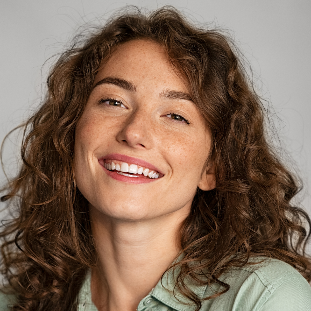 Close-up of a woman with curly red hair smiling and showing teeth, wearing a light green shirt.