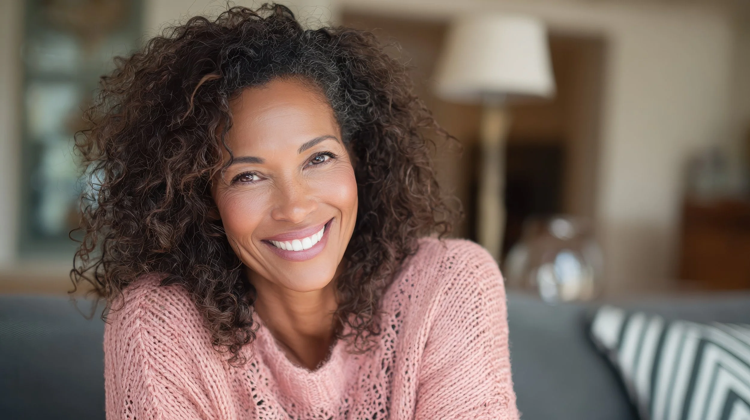 A woman with curly hair smiling and wearing a pink sweater, sitting on a couch in a cozy living room.