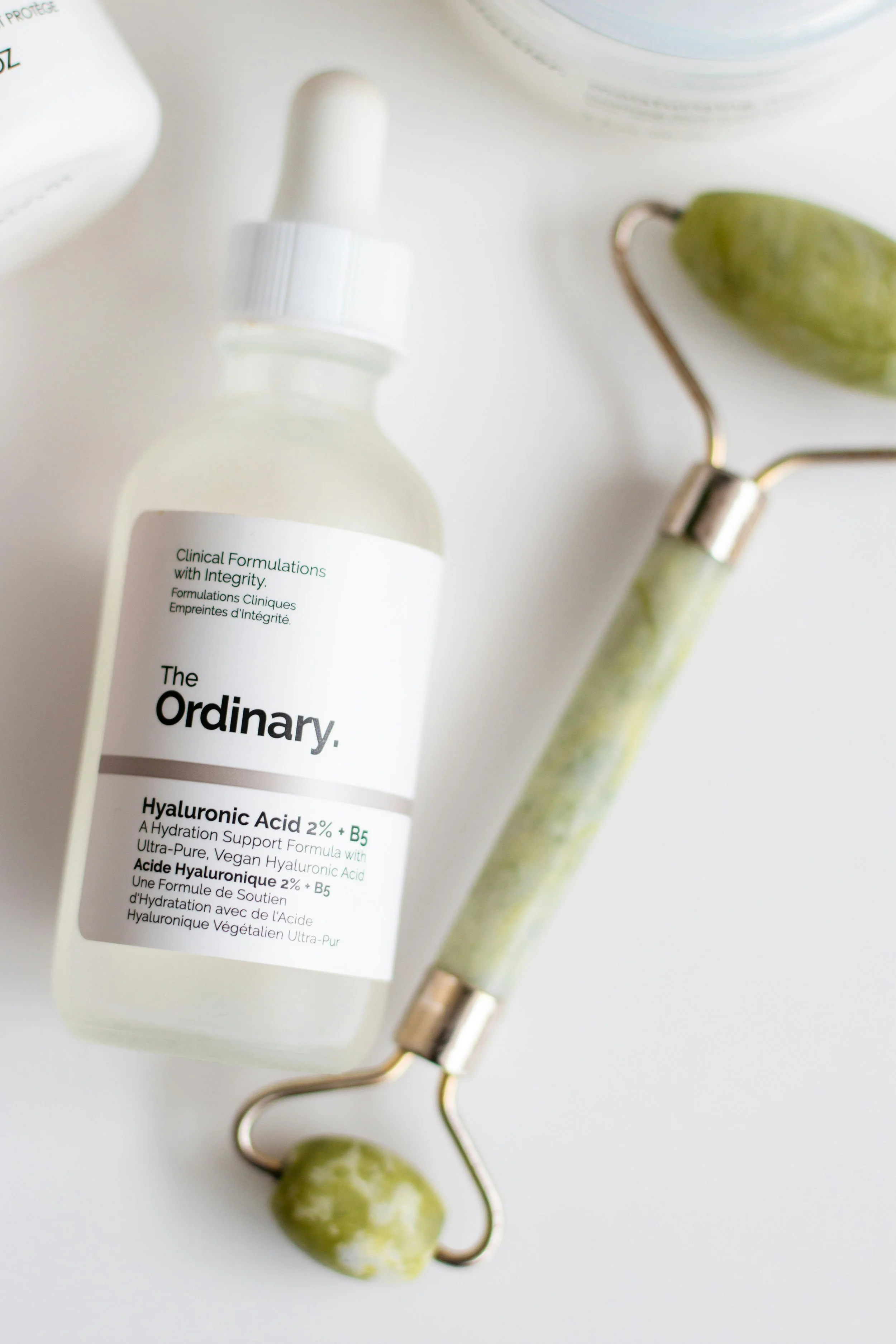A bottle of The Ordinary Hyaluronic Acid 2% + B5 serum, a jade roller, and a jade gua sha on a white surface.