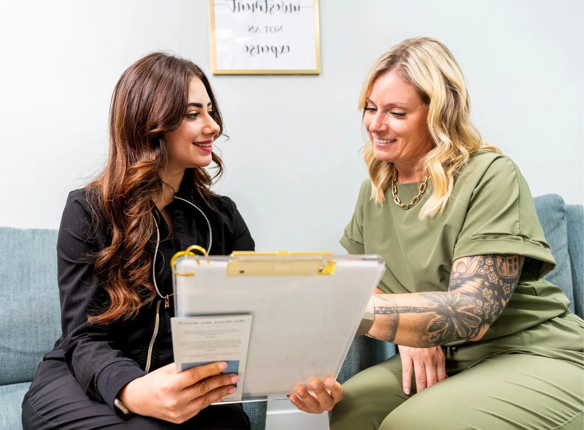 A young woman in a black jacket sitting on a couch with a stethoscope around her neck, speaking with a woman in green scrubs with tattoos on her arm, holding a clipboard in a medical office.