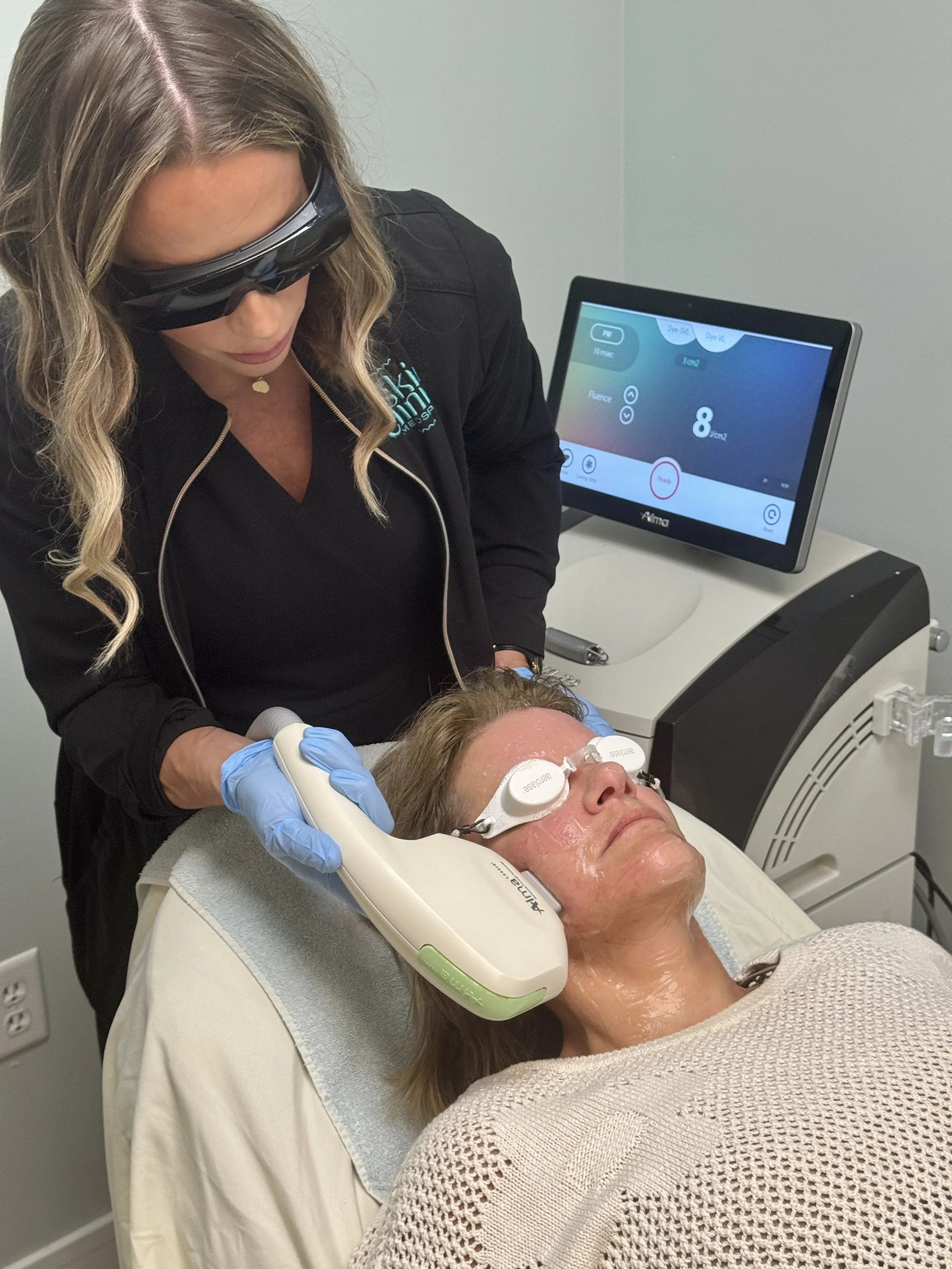 A woman receiving a facial treatment with a handheld device while lying on a treatment bed, wearing protective glasses, with a technician operating the device in a clinical setting.