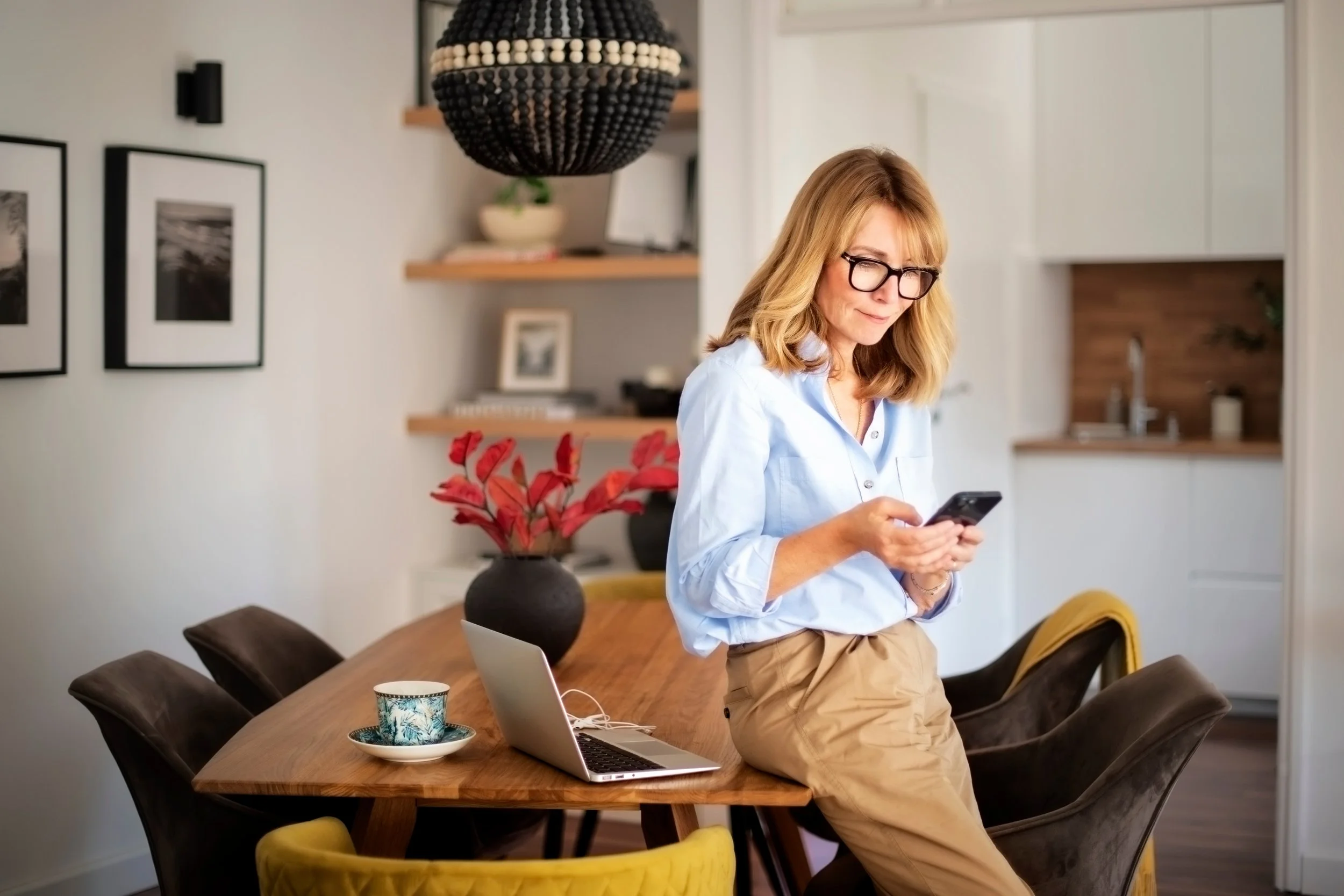 A woman with shoulder-length blonde hair and glasses is standing in a dining area, looking at her phone. She is wearing a light blue shirt and beige pants. The dining table has a laptop, a cup with a saucer, and a black vase with red leaves. The background shows wall art, a bookshelf, and a kitchen with white cabinets and a wooden backsplash.