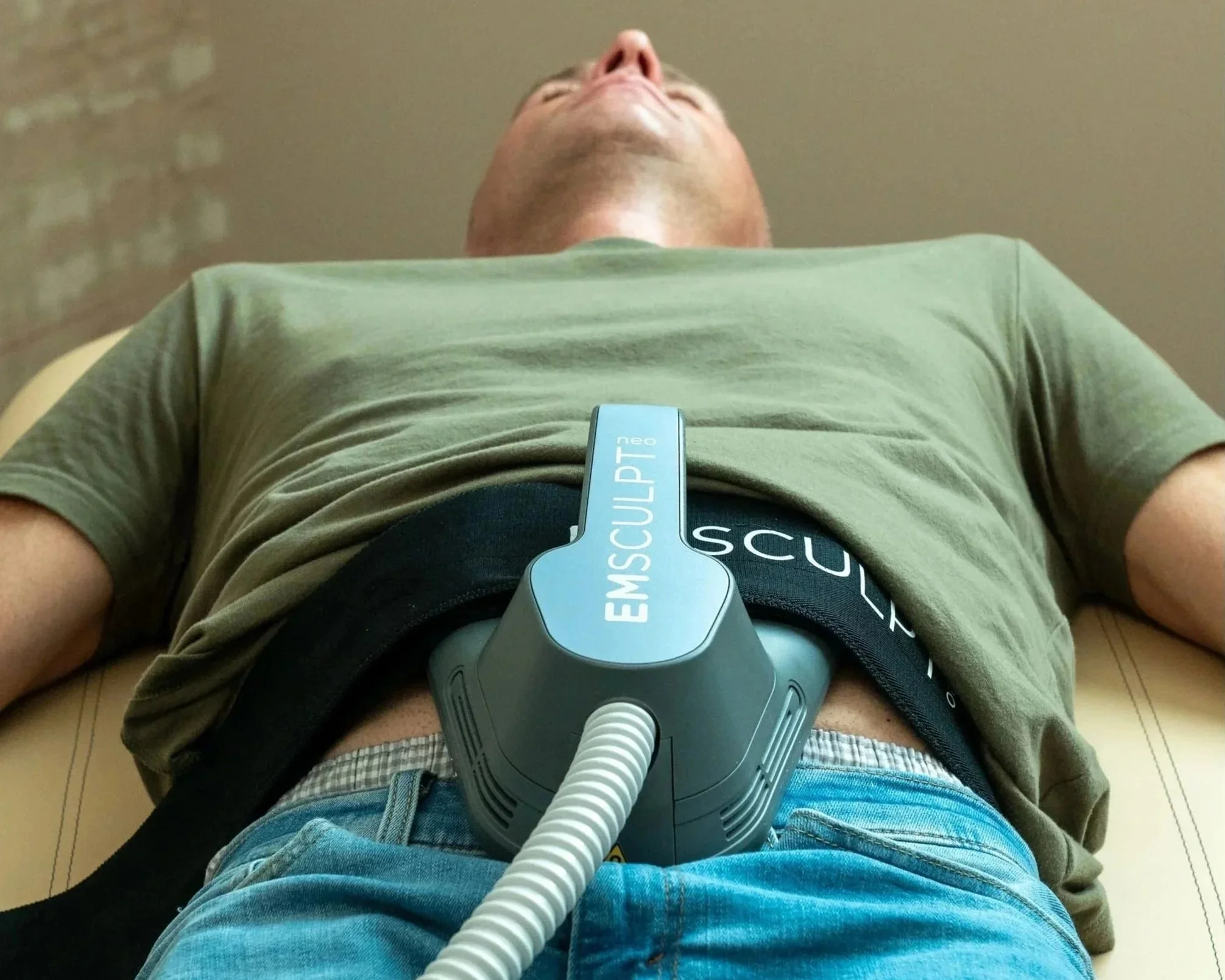 A person lying on a medical examination table with a pulmonary device attached to their chest, wearing a green shirt, blue jeans, and plaid underwear, with a beige tiled floor behind them.