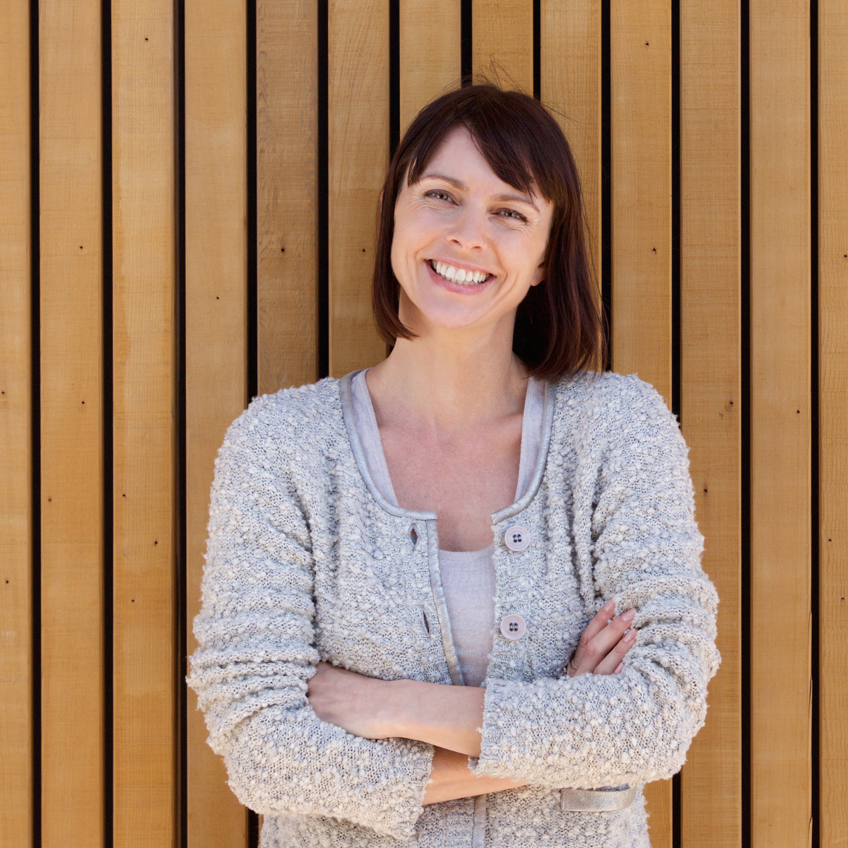 A woman with shoulder-length dark brown hair smiling, standing with arms crossed in front of a wooden slatted background.