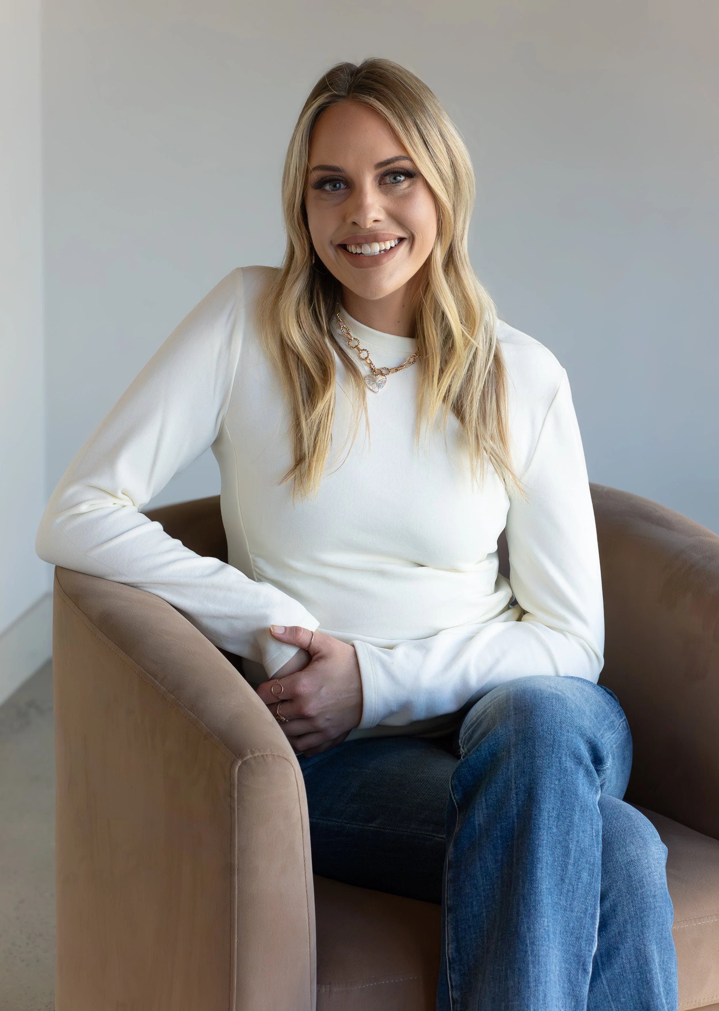 Portrait of a young woman with blonde hair, blue eyes, smiling, sitting in a beige armchair, wearing a white long-sleeve top and blue jeans, in a room with plain white walls.