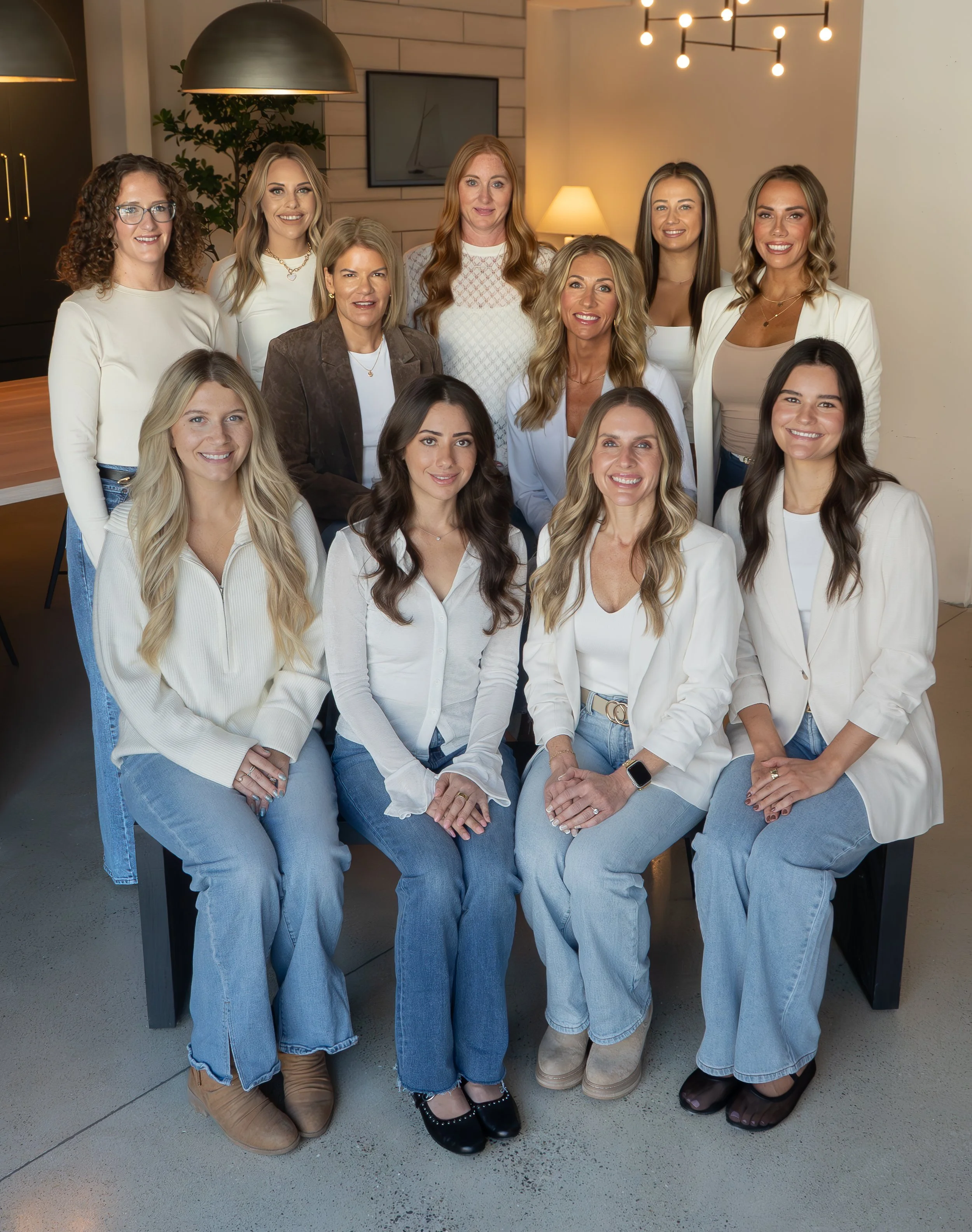 Group portrait of fifteen women indoors, some seated and some standing, in a modern, well-lit space with a wooden table and decorative lighting.