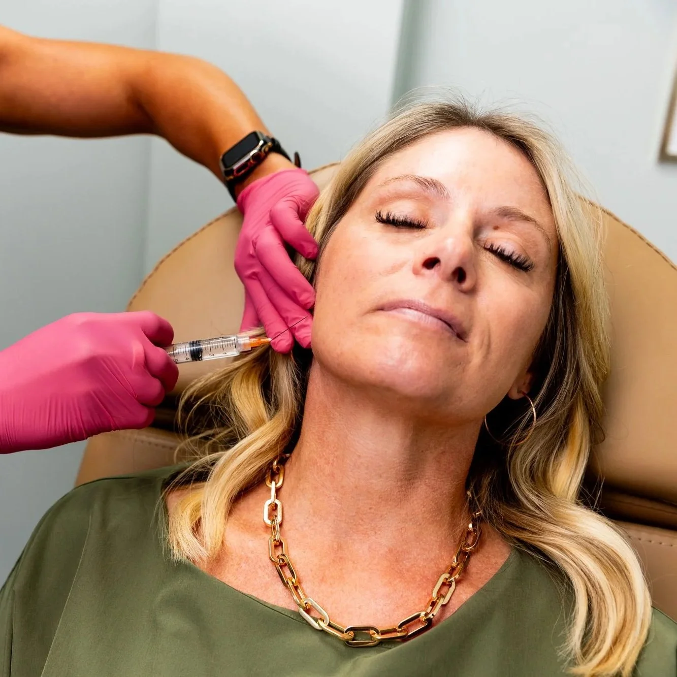 A woman receiving a cosmetic injection in her face from a professional wearing pink gloves at a medical clinic.