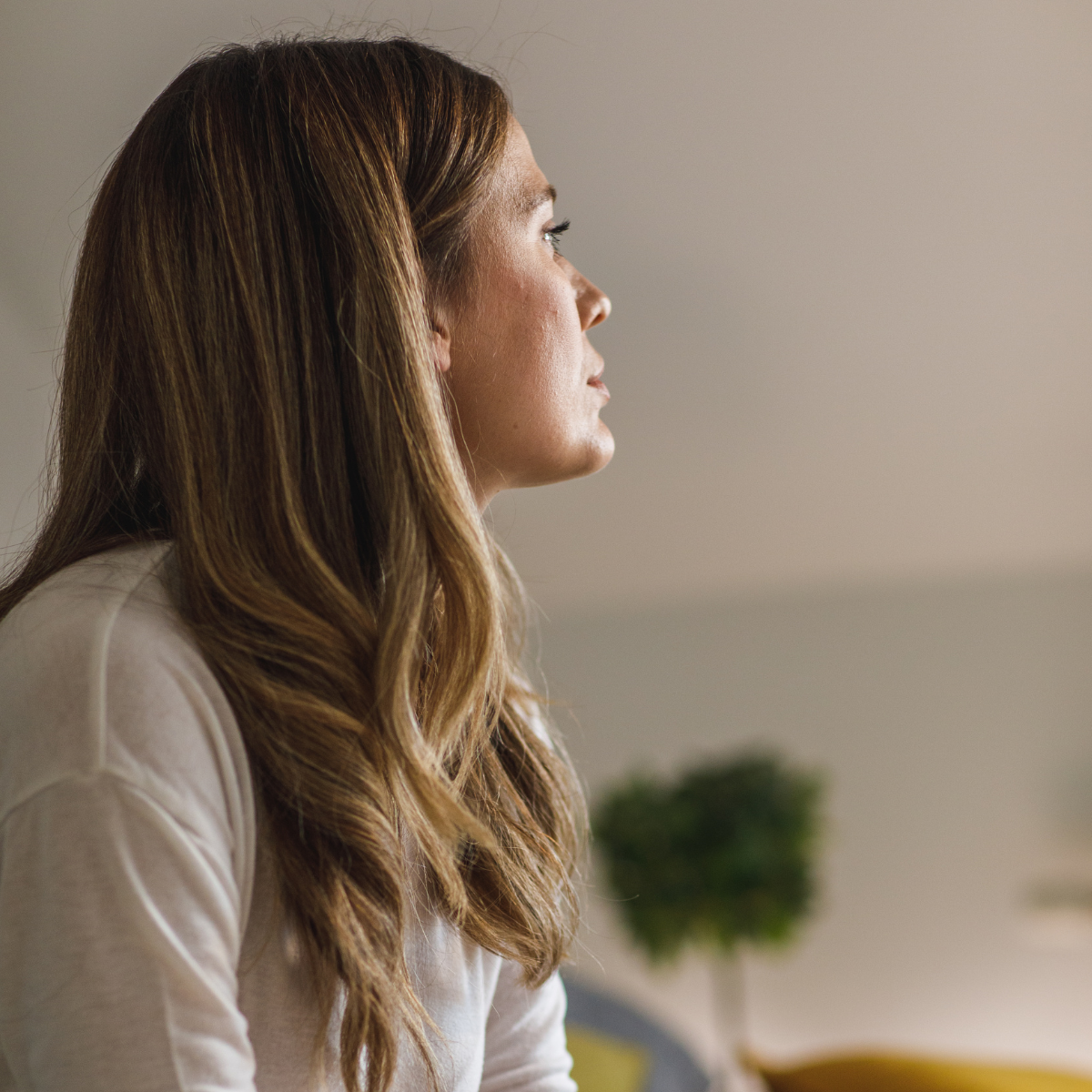 Profile of a woman with long, wavy brown hair looking to the right, sitting indoors with a blurred background.