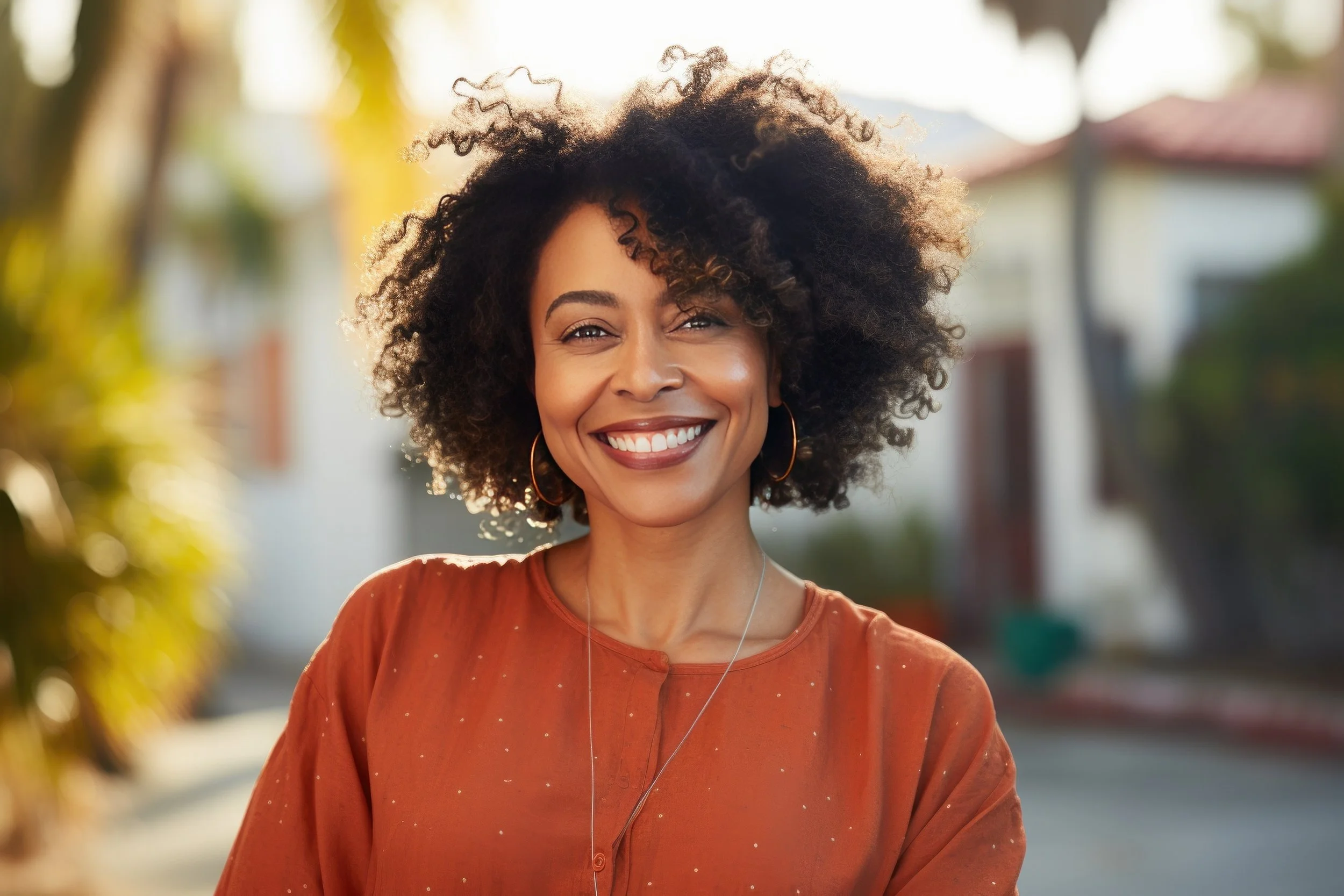 A woman with curly hair smiling outdoors during daytime, wearing an orange top and hoop earrings.