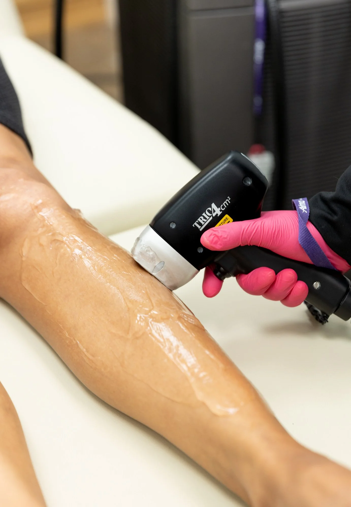 A person receiving a medical treatment on their leg using a handheld device, with a healthcare worker wearing pink gloves.