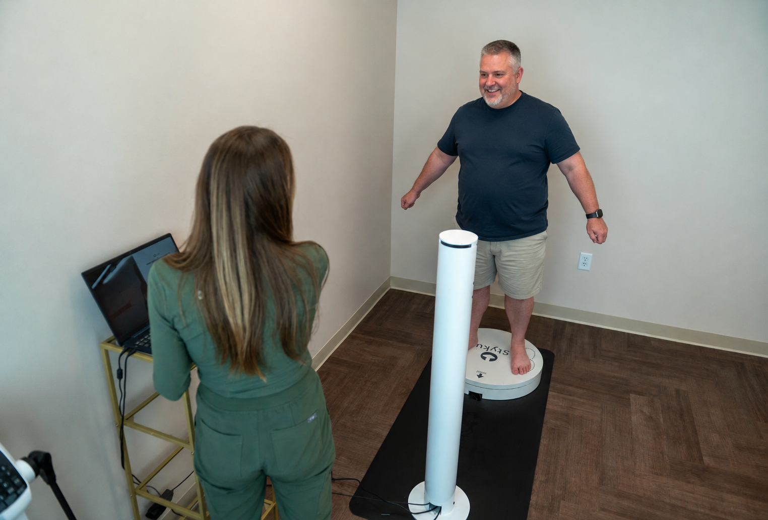 A man standing barefoot on a body composition scale in a room, smiling, with a woman in medical scrubs checking the scale's display on a laptop.