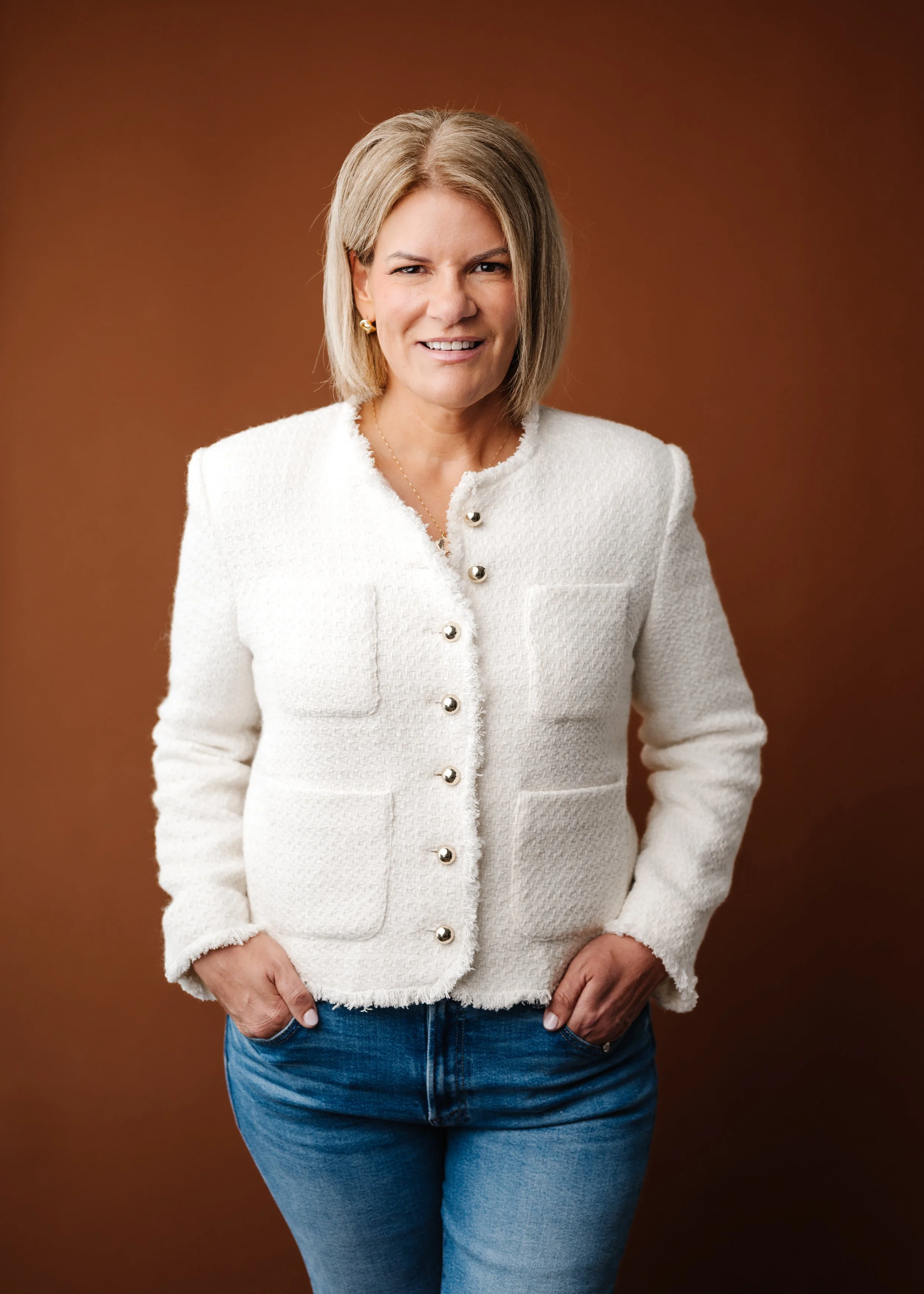 A woman with blonde hair, wearing a white textured jacket with pearl buttons and blue jeans, standing against a brown background.