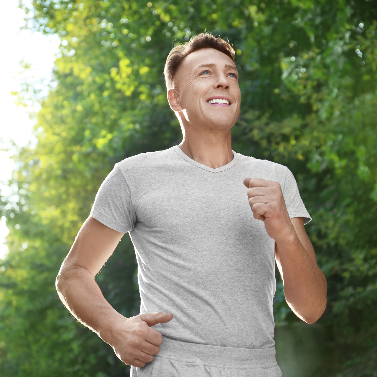 A middle-aged man running outdoors on a sunny day with green trees in the background, wearing a light gray t-shirt, smiling, and looking upward.