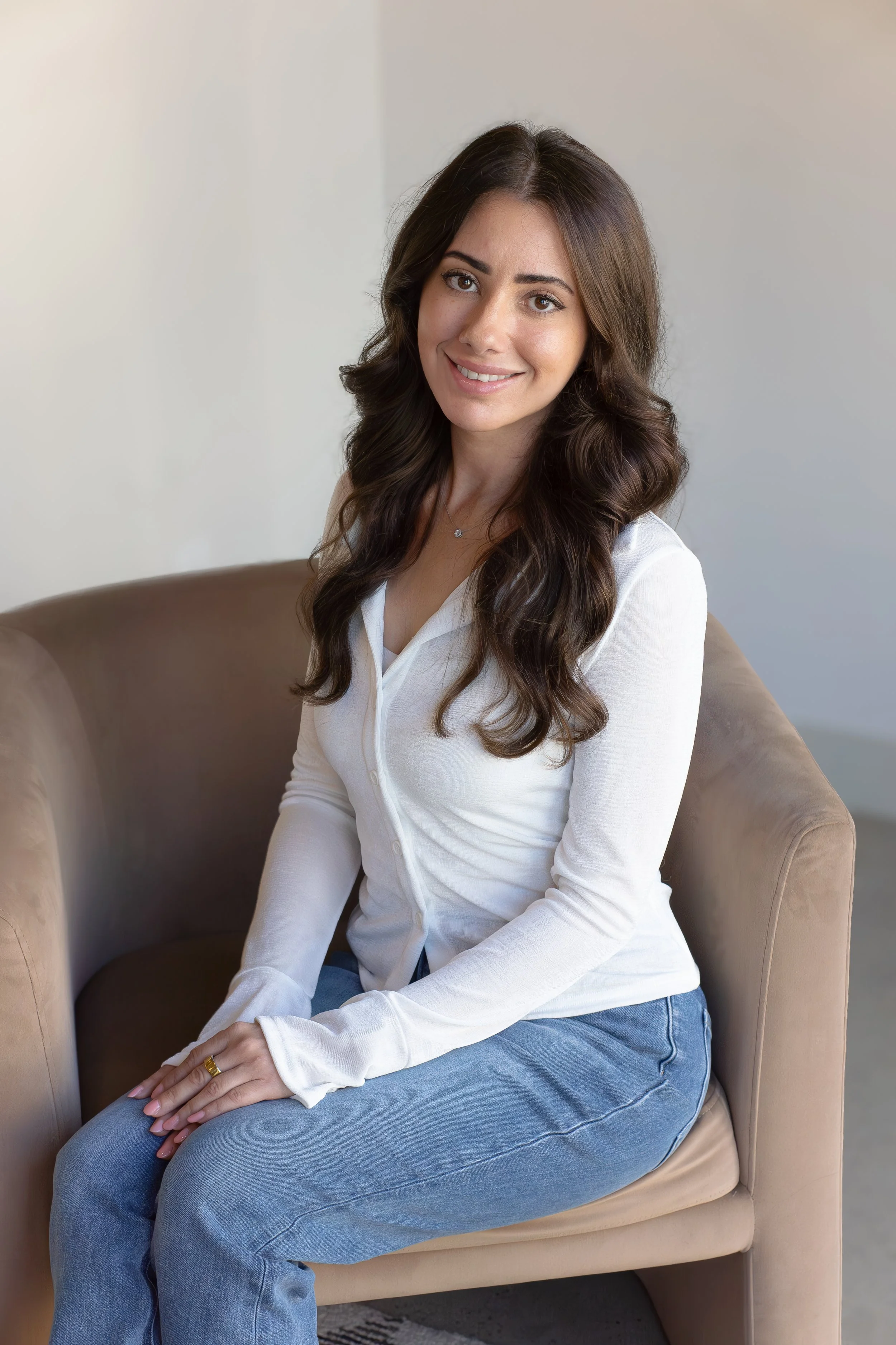 A woman with long brown hair, wearing a white blouse and blue jeans, sitting on a beige chair and smiling at the camera.