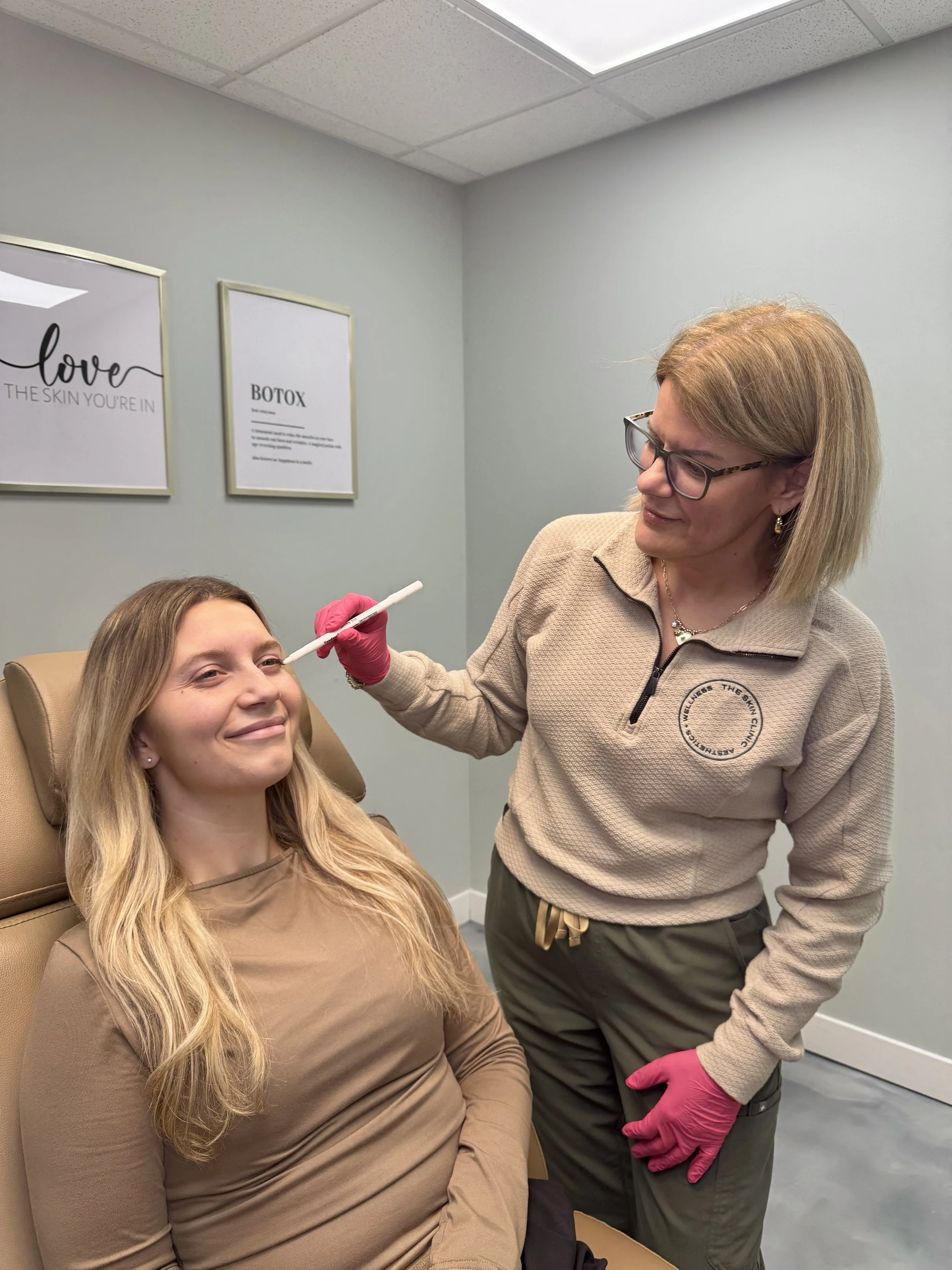 A woman with long blonde hair lying in a medical chair, smiling, as a healthcare professional prepares to administer a Botox injection near her forehead.
