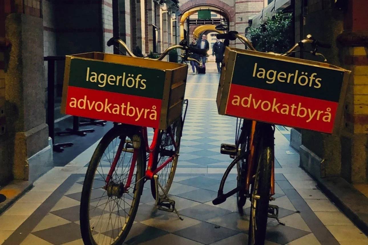 Två cyklar med reklamskyltar som säger "lagerlöfs advokatbyrå" står på en gångbana i en stadsgata. I bakgrunden syns människor