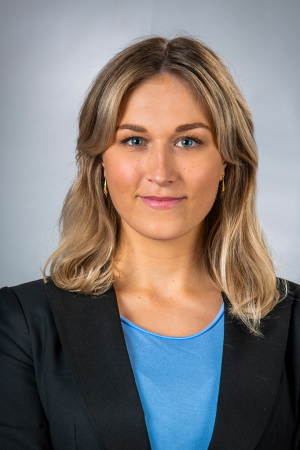 Ung kvinna med blont, vågigt hår, blå ögon, vit skjorta och svart blazer mot en ljusgrå bakgrund.