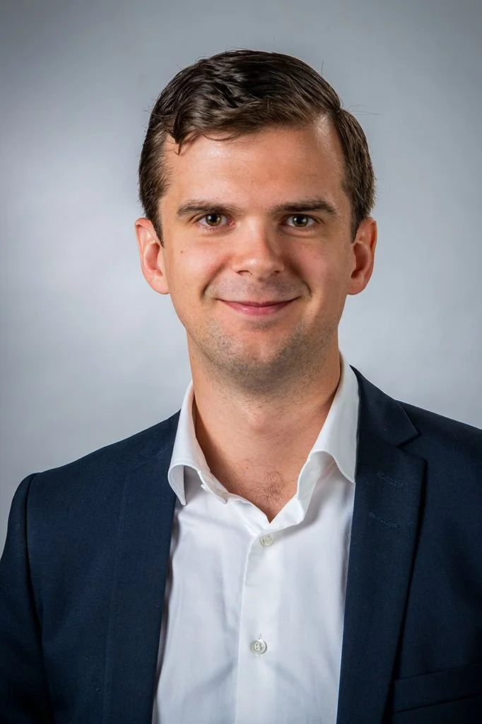 Ung man i affärskostym och vit skjorta, ler mot kameran, mot en grå bakgrund.