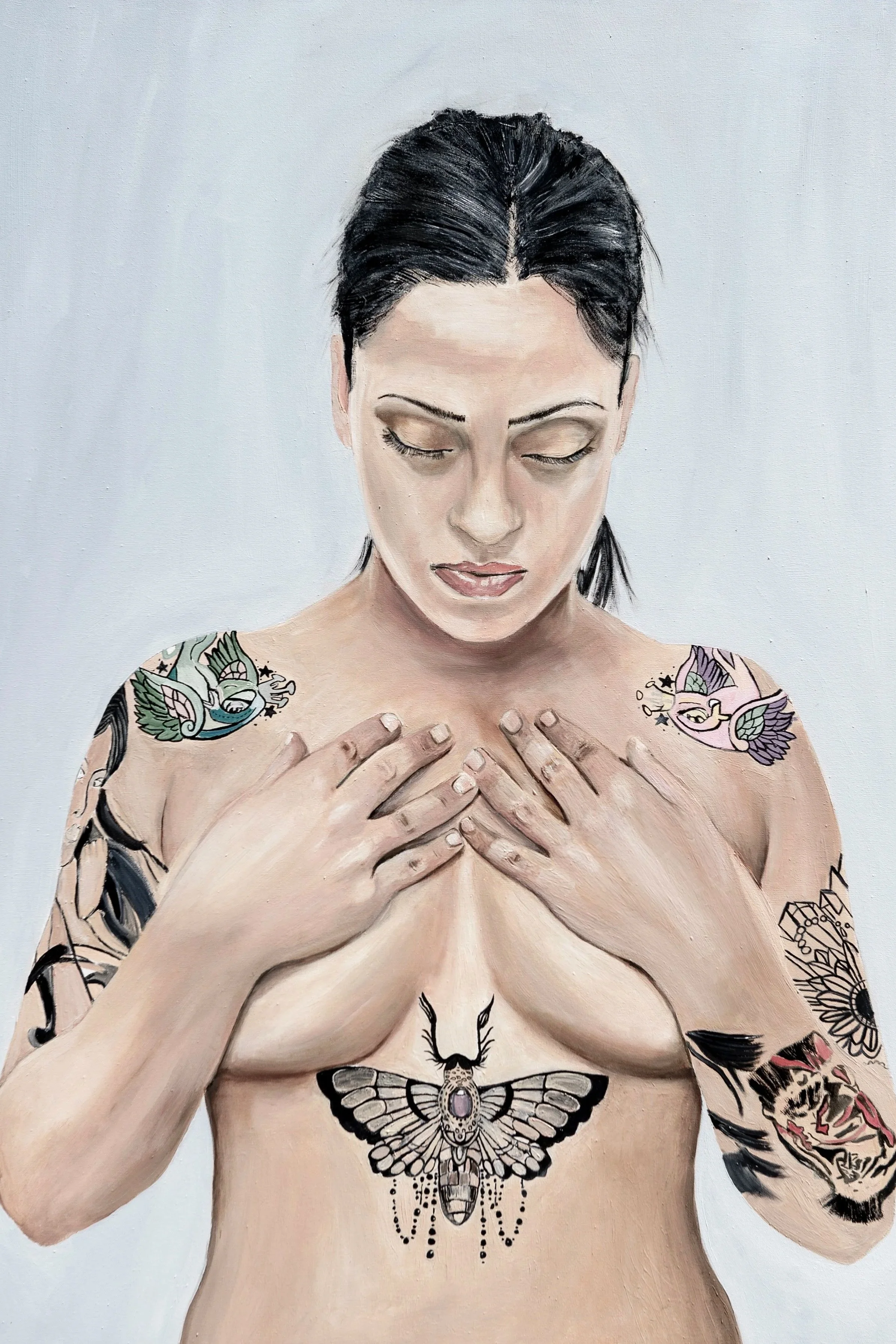 A woman with short black hair and multiple colorful tattoos, including a moth on her chest, is topless with her hands crossed over her chest, looking down with closed eyes.