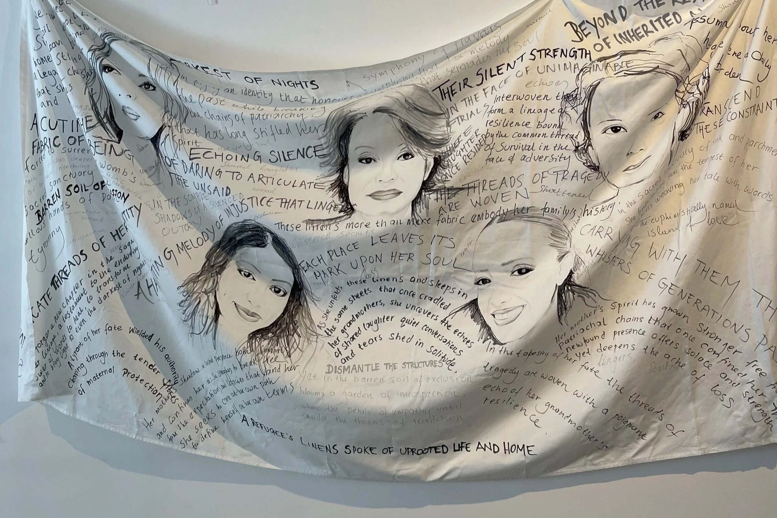 A white fabric artwork featuring five illustrated female faces with handwritten poetic text surrounding them.