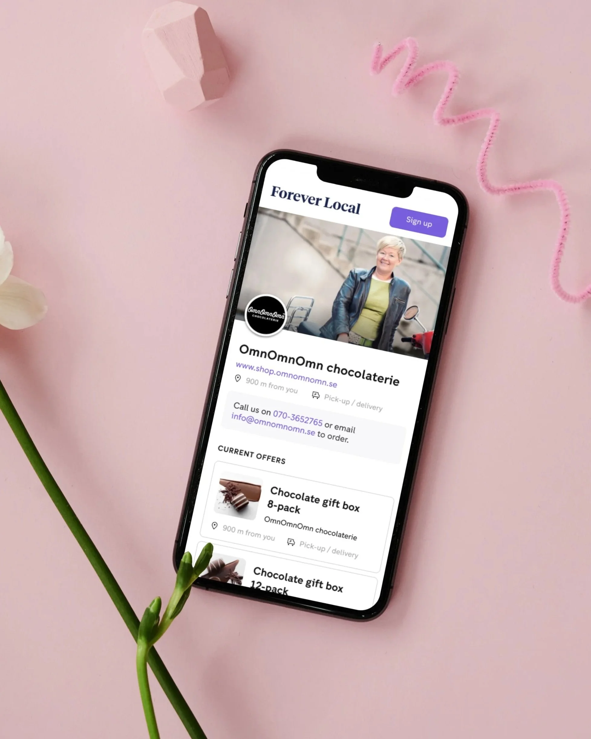 Smartphone displaying a website for a local chocolatier called OmOmOmn chocolate shop, showing contact information and special offers for chocolate gift boxes, placed on a pink surface with decorative pink pipe cleaner, a pale pink geometric object, and a white flower with green stem in the surroundings.