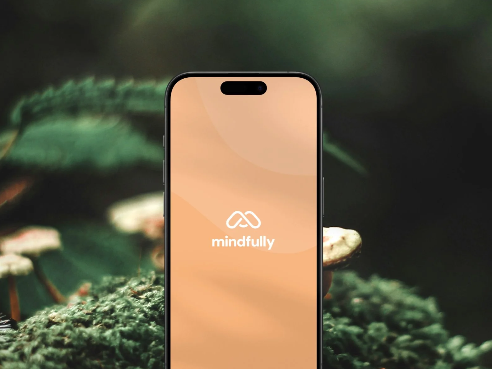 A smartphone displaying the logo and name 'mindfully' on its screen, held in front of a blurred background of a green mossy surface with small mushrooms.