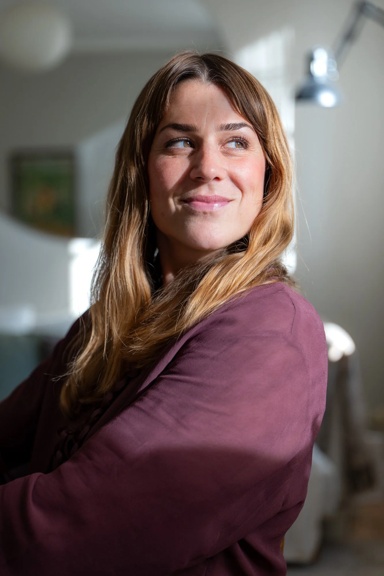 A woman with long, wavy, light brown hair and blue eyes, looking to her left with a slight smile, wearing a maroon blazer, indoors with a blurred background.