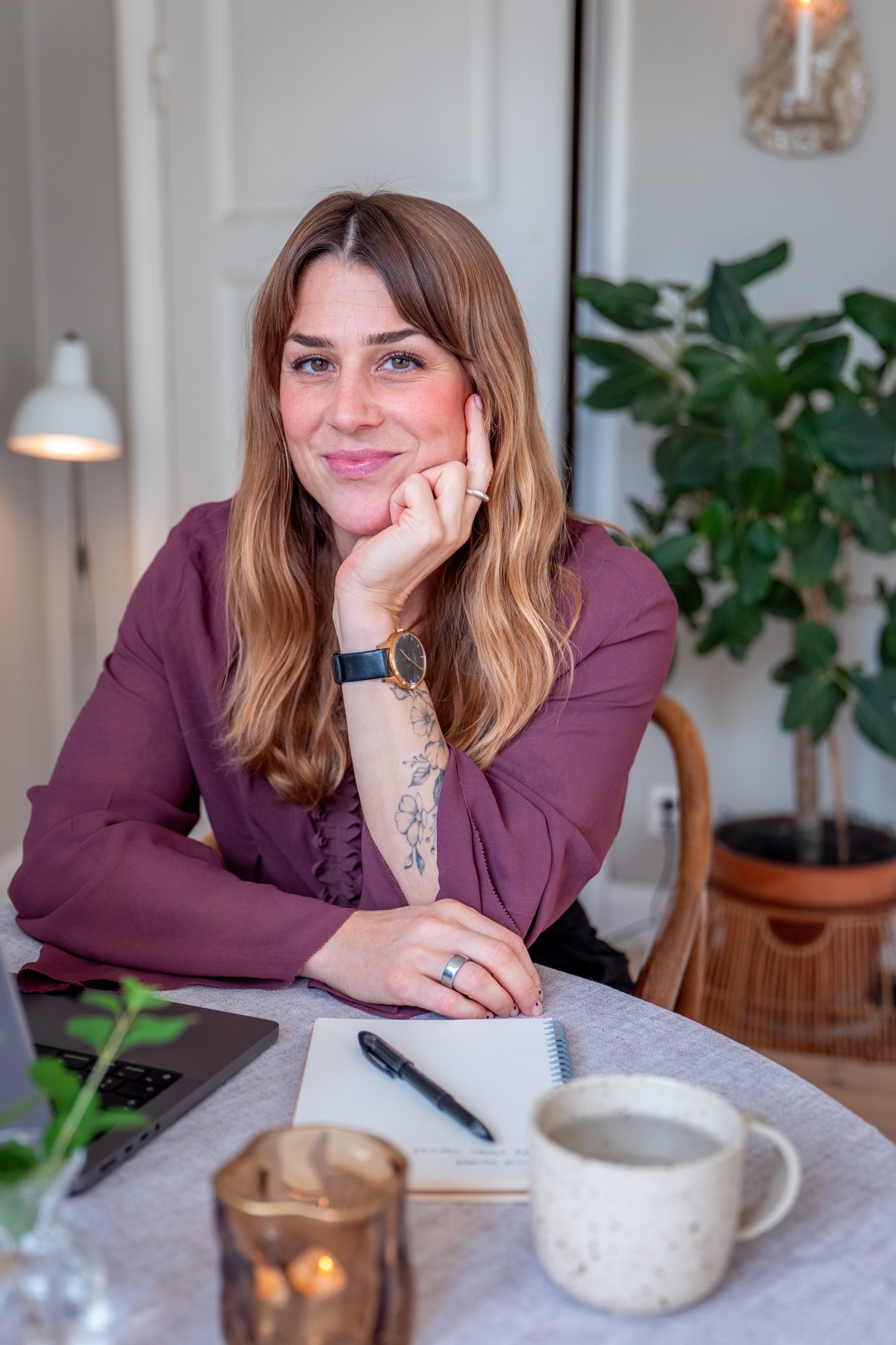 A woman with light skin, long wavy hair, wearing a maroon blouse, seated at a table with a notebook, pen, cup, and a laptop, smiling and resting her chin on her hand in a cozy, well-lit room with a plant and lamp in the background.