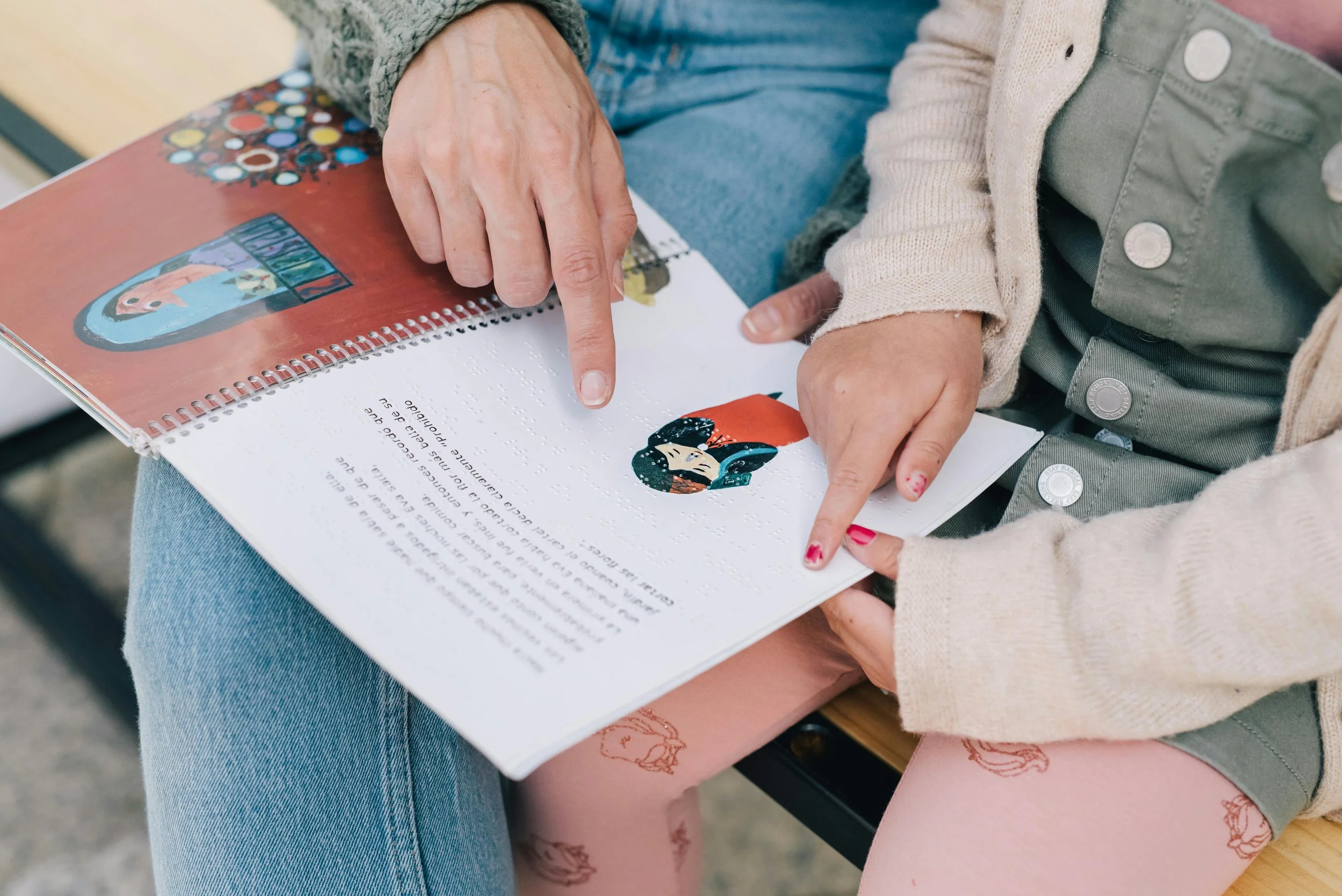 Deux personnes regardent un livre pour enfants, l'une pointe une illustration de personnage, assises côte à côte, avec un fond flou.