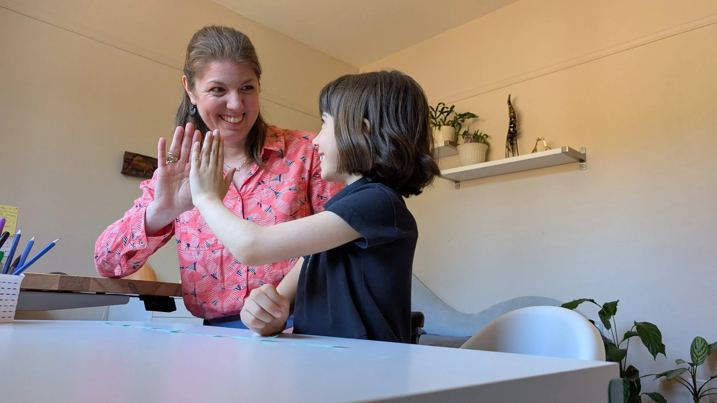 Une femme souriante échange un high five avec une jeune fille assise à une table dans une pièce bien éclairée.