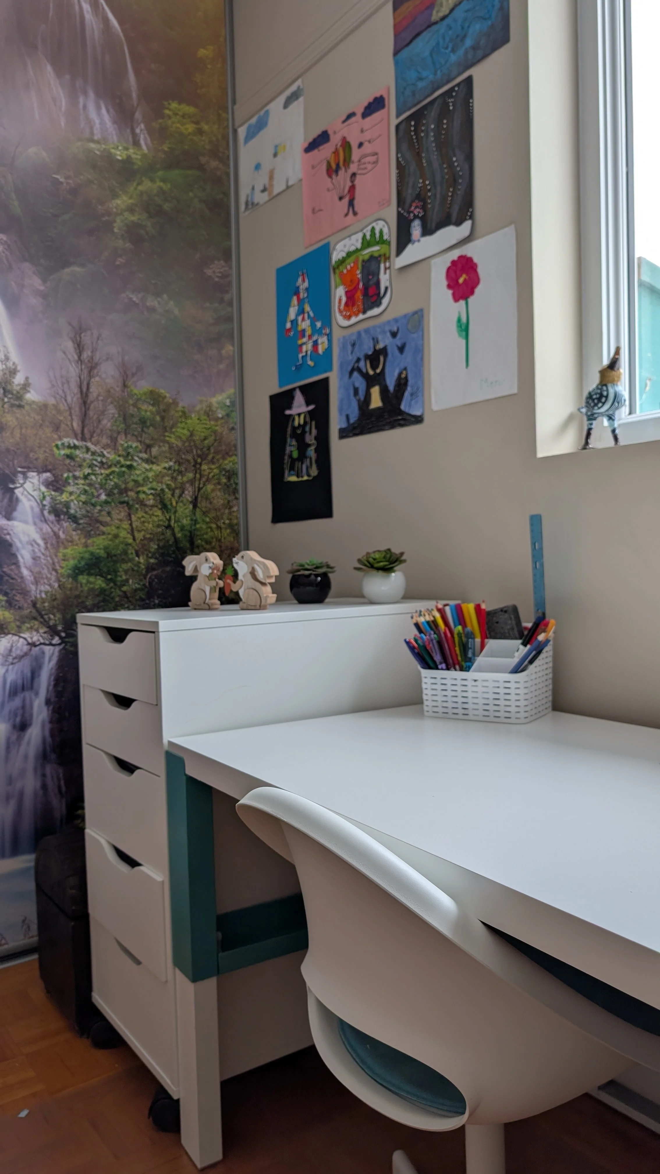Un bureau blanc avec une chaise blanche, des stylos colorés dans un panier, une étagère à tiroirs, décorations en forme de lapins, et des dessins d'enfants sur le mur, avec un fond de forêt tropicale.
