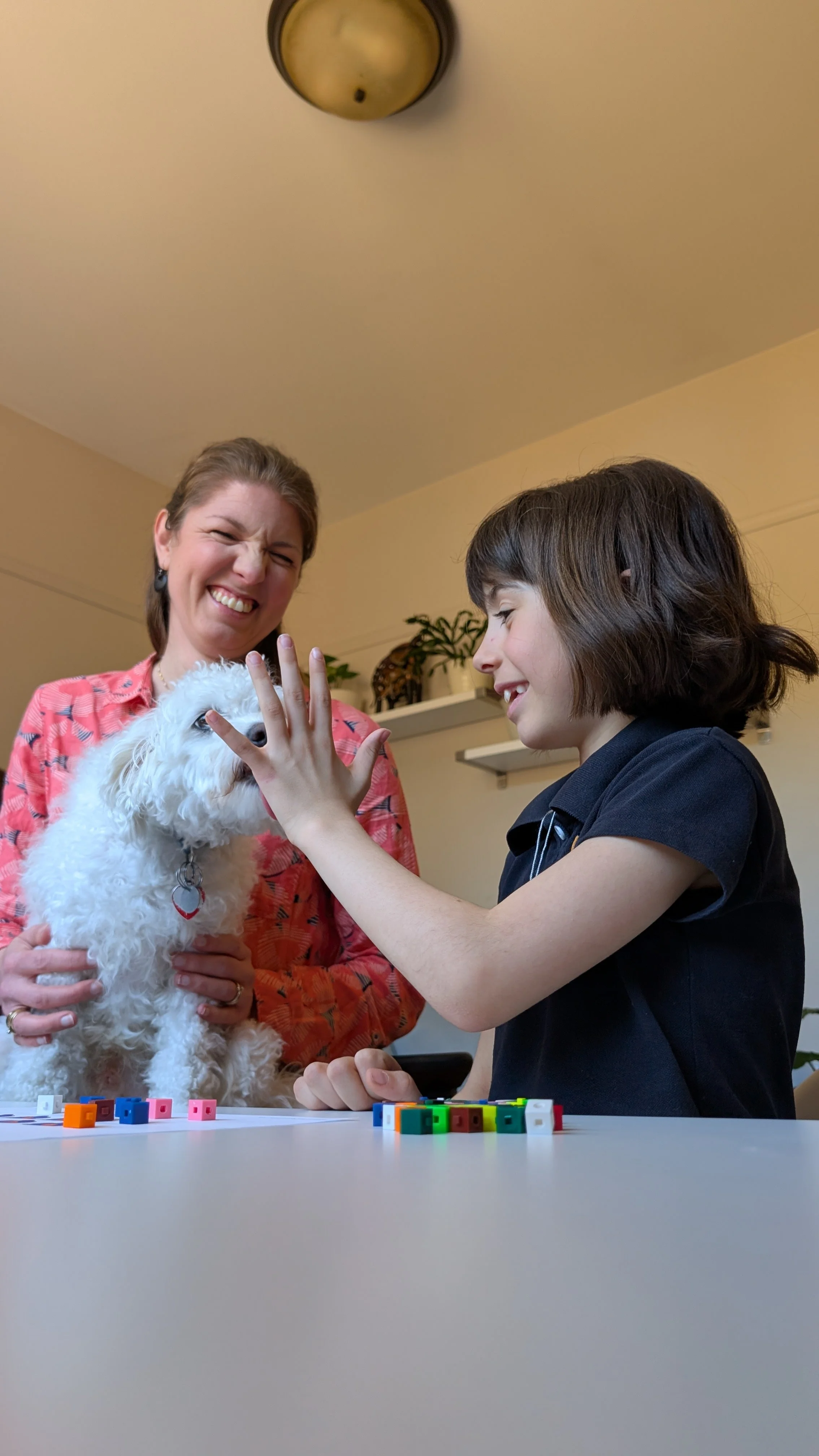 Une femme et une jeune fille jouent avec un chien blanc dans une pièce intérieure. La femme porte une chemise rose, la fille porte une chemise noire. Il y a des petits blocs colorés sur la table devant eux.