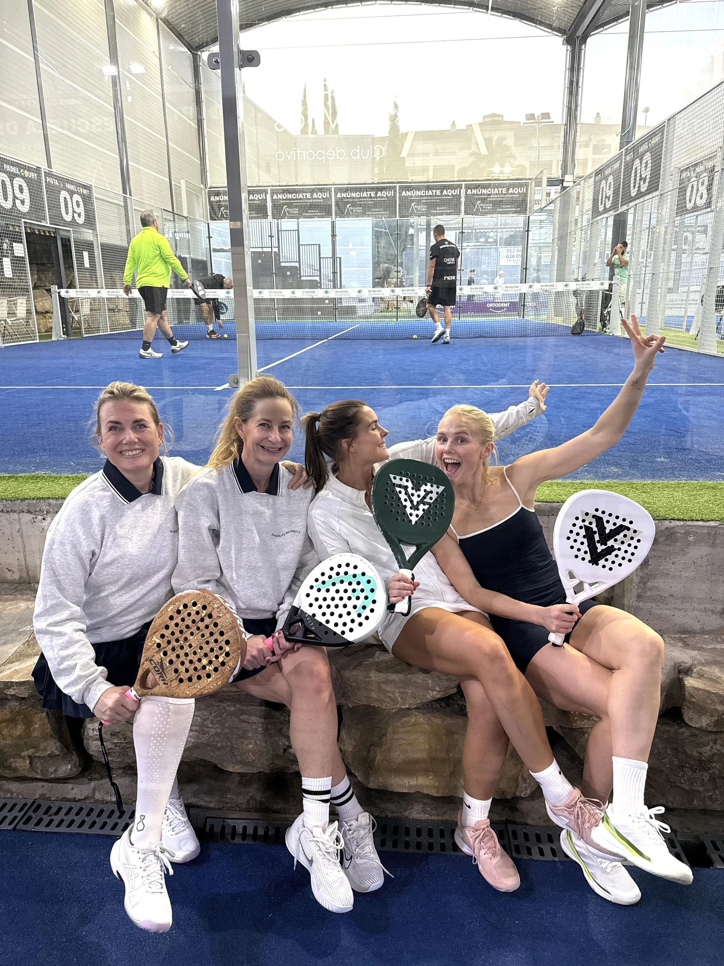 Four women sitting on a stone ledge, holding pickleball paddles, smiling at the camera, with a pickleball court in the background.